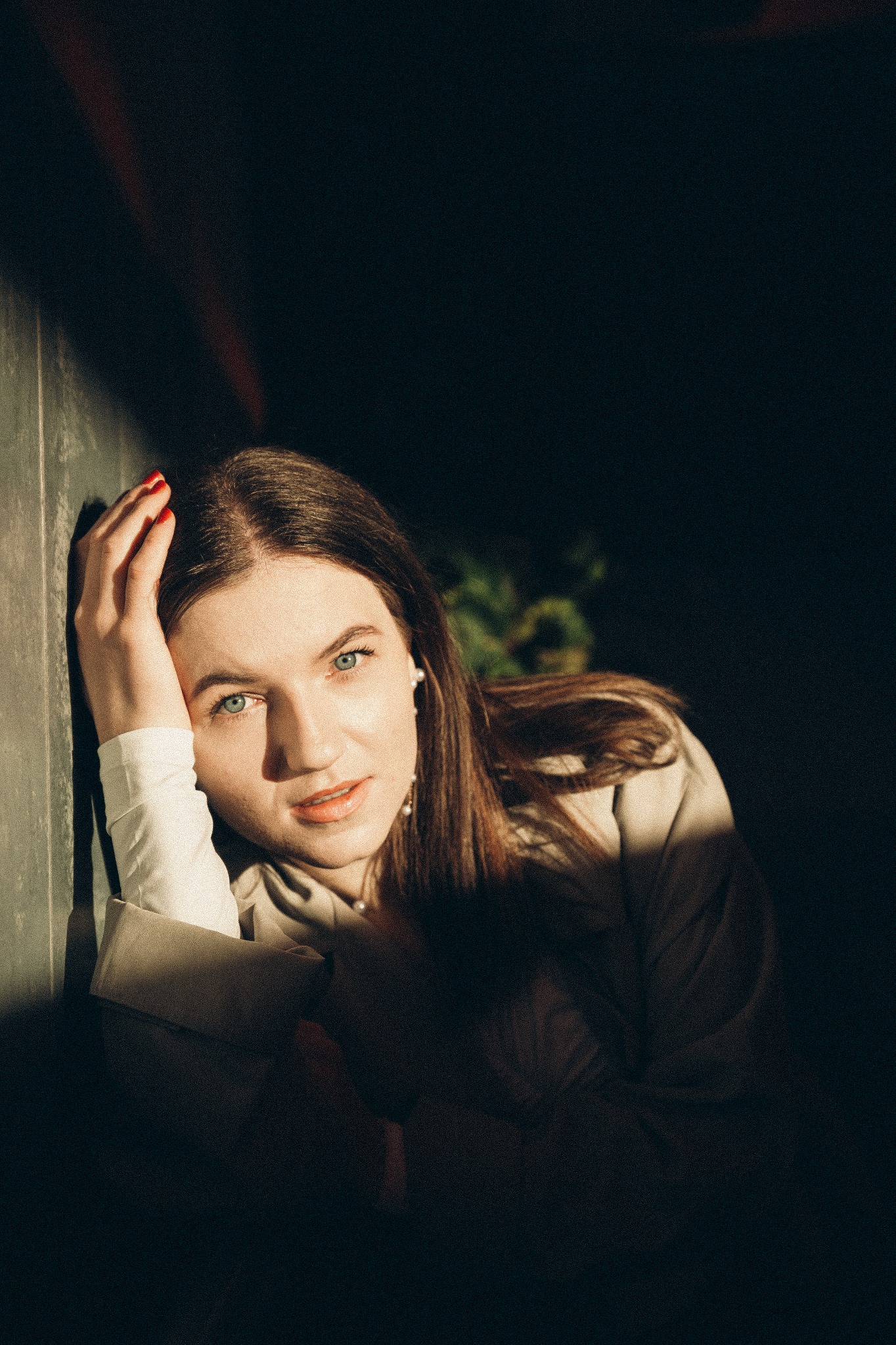 Portret młodej kobiety z niebieskimi oczami, opartej o ścianę w mocnym kontraście światła i cienia, z widocznym czerwonym manicure.