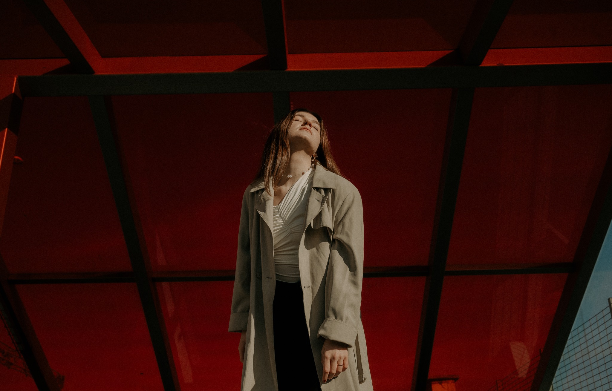 Kobieta w płaszczu pozuje z zamkniętymi oczami pod czerwoną konstrukcją, oświetlona promieniami słońca.