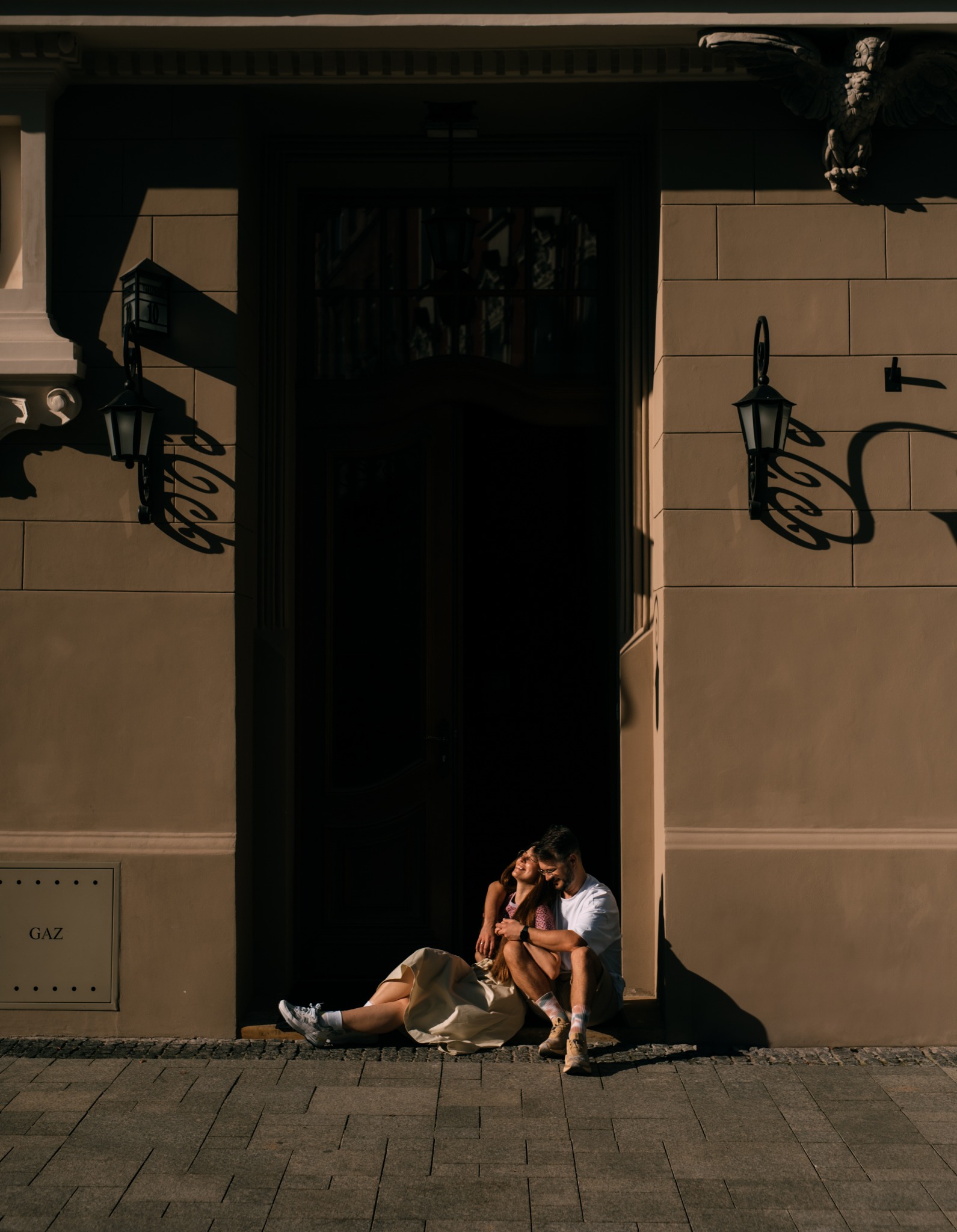 Uśmiechnięta para siedzi na chodniku przed budynkiem w promieniach słonecznych, kobieta w sukience i sportowych butach, mężczyzna w białej koszulce i krótkich spodenkach.