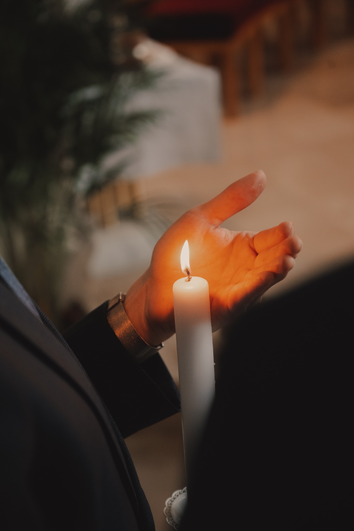 Dłoń mężczyzny w garniturze osłania płomień zapalonej, białej świecy podczas uroczystości, widoczny złoty zegarek na nadgarstku.