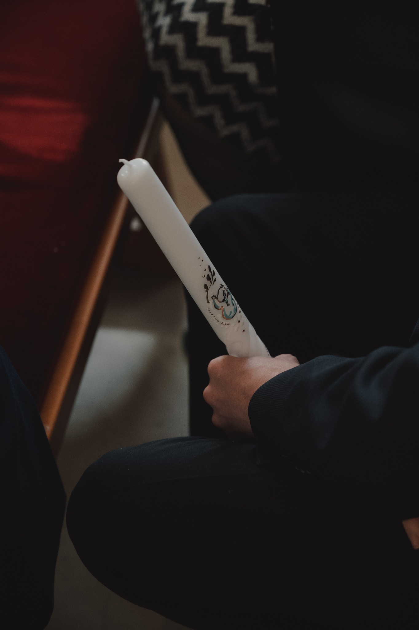 Dłoń trzyma białą, zdobioną świecę z motywem roślinnym i aniołka, prawdopodobnie podczas uroczystości religijnej.