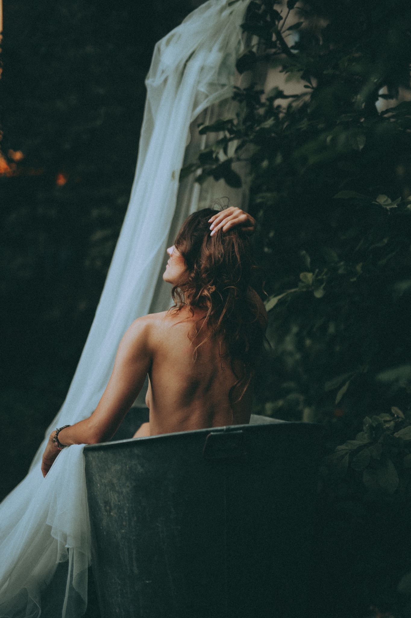 Kobieta siedząca w metalowej wannie na tle zieleni, z delikatnym białym materiałem zwisającym za nią, głowę opiera na dłoni, w tle ciemne liście i gałęzie.