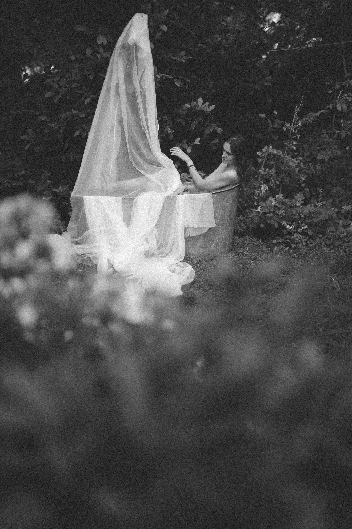 Kobieta w wannie z baldachimem z tiulu na tle zieleni, stylizowana sesja w plenerze, czarno-biała fotografia.