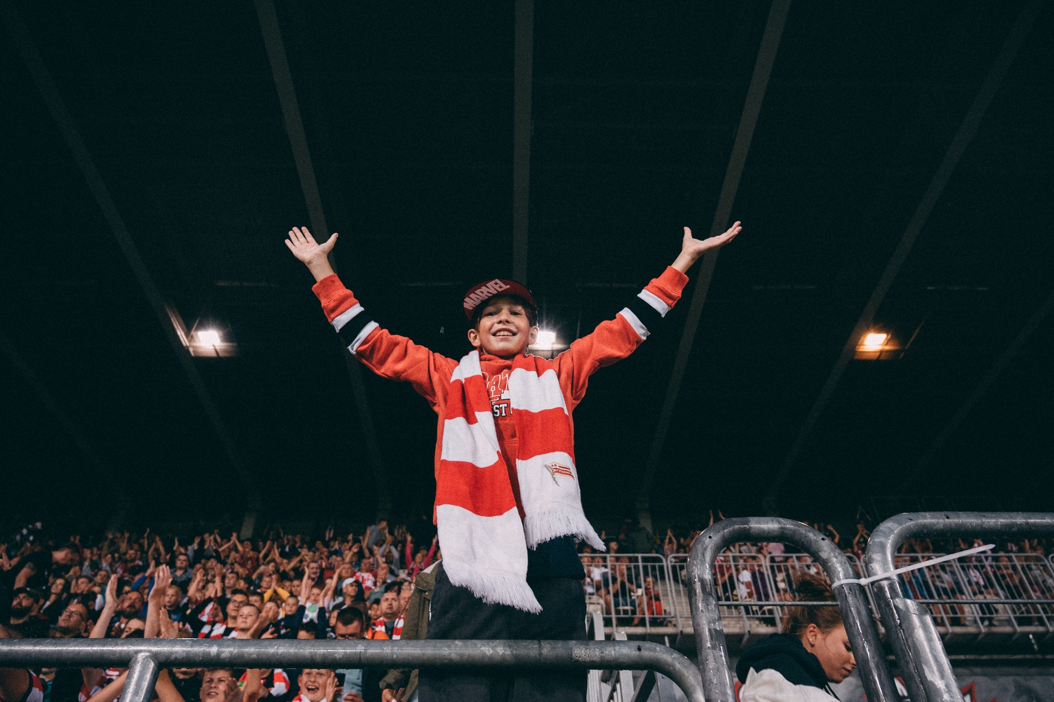 Uśmiechnięty chłopiec w czerwono-białym szaliku i czapce z daszkiem, z rozłożonymi rękami, świętuje na trybunach stadionu podczas wydarzenia sportowego, z tłumem kibiców w tle.