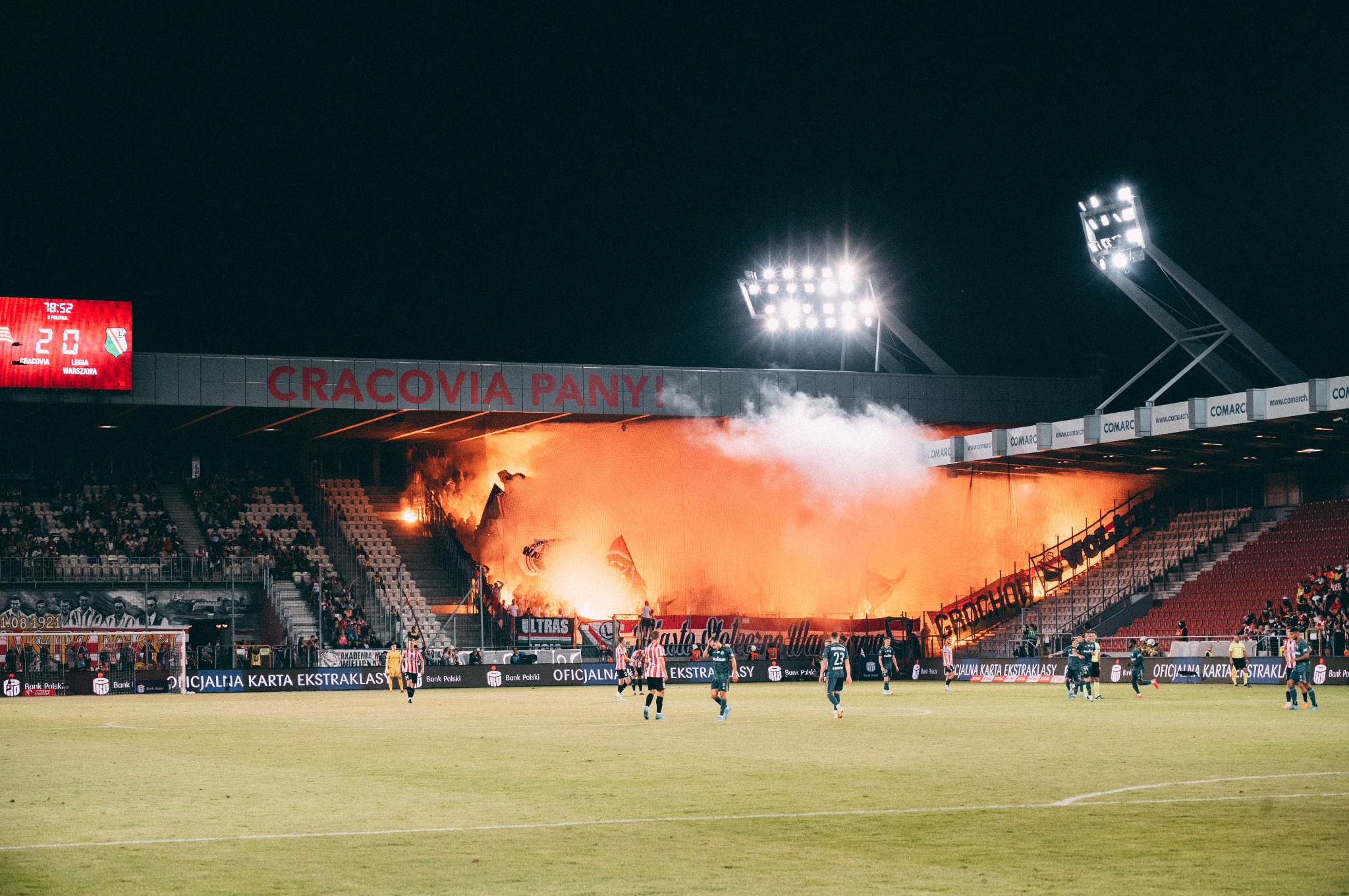 Stadion piłkarski podczas meczu, kibice odpalają race, tworząc pomarańczową zasłonę dymu na trybunach, widoczni piłkarze na boisku i tablica wyników.