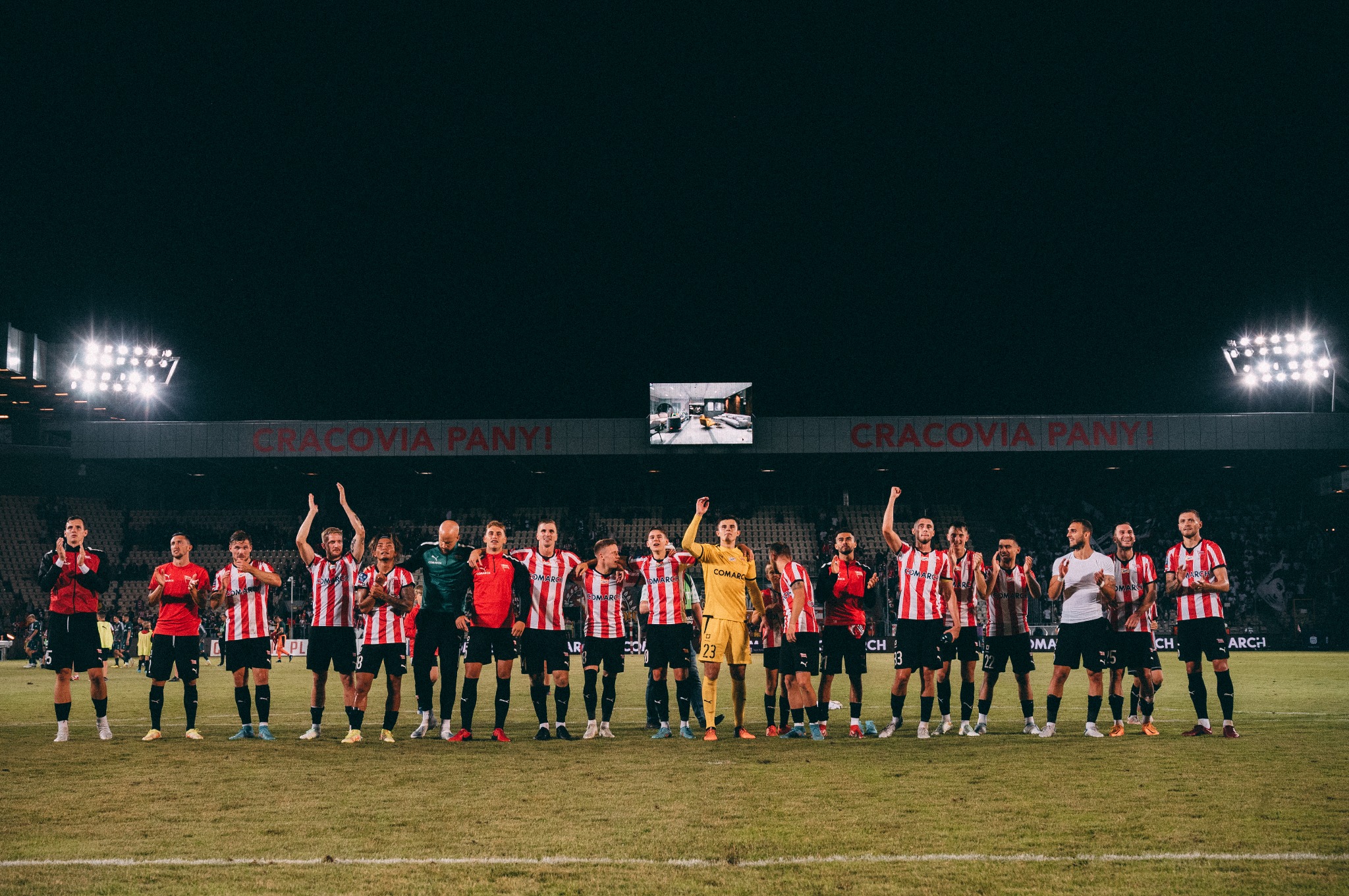 Drużyna piłkarska Cracovii świętuje zwycięstwo na stadionie, oświetlonym jupiterami, z widocznym telebimem nad trybunami.