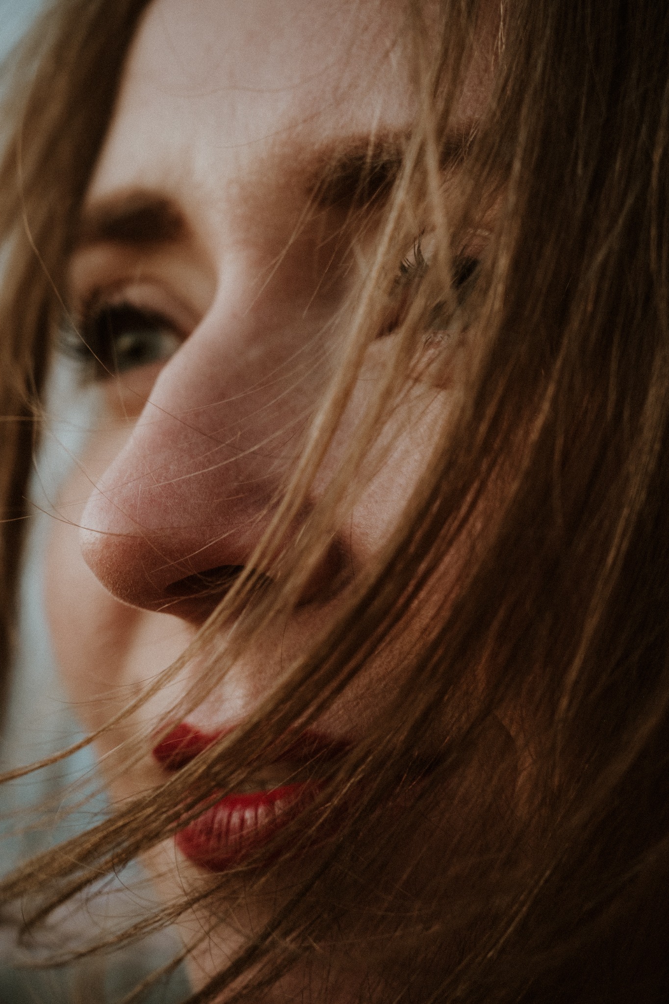 Zbliżenie na profil kobiety z rozpuszczonymi, jasnymi włosami częściowo zasłaniającymi twarz, z widocznym zielonym okiem i czerwonymi ustami.