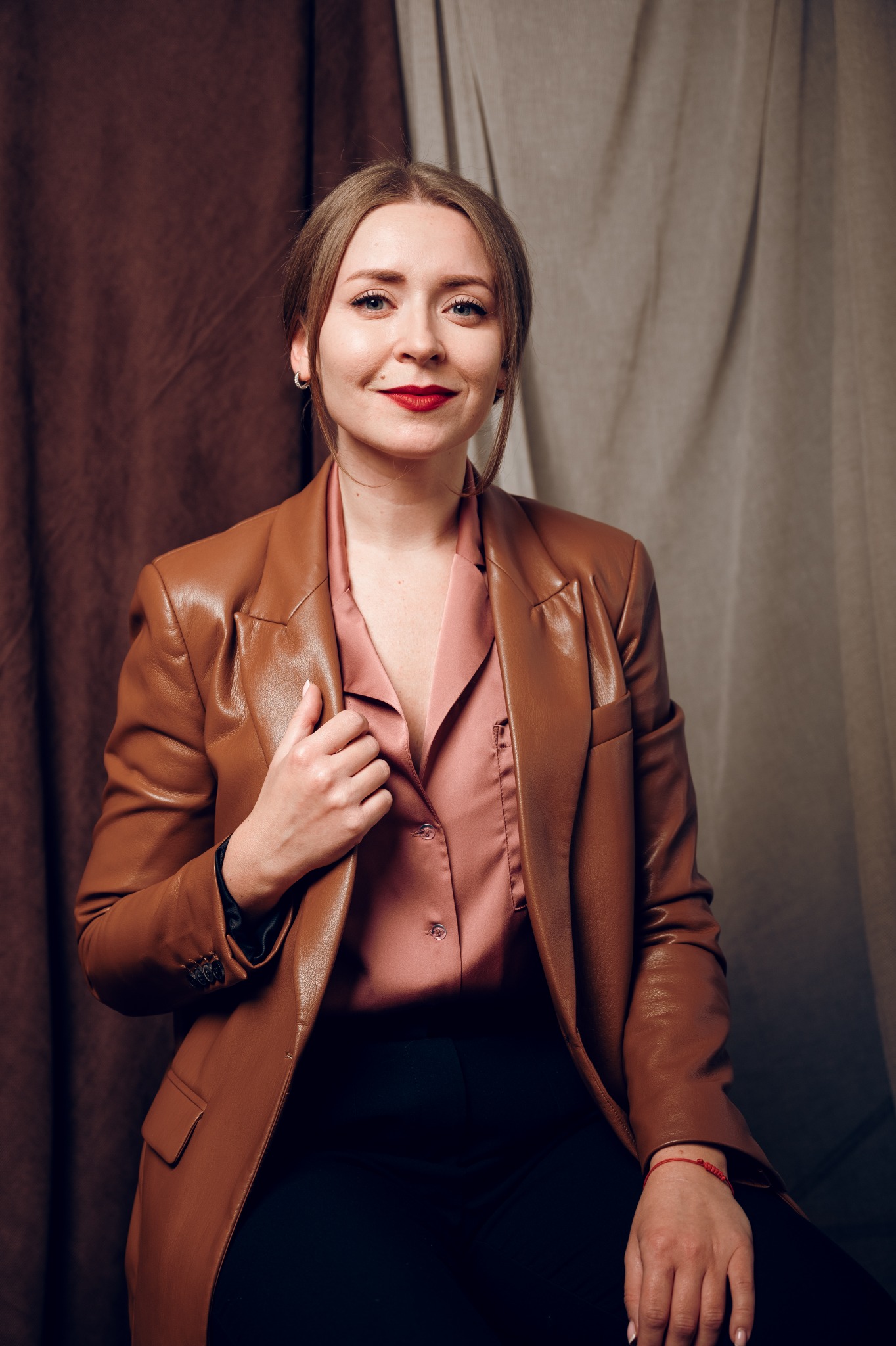 Portret kobiety w brązowej skórzanej marynarce i różowej koszuli, z delikatnym uśmiechem i czerwonymi ustami, na tle drapowanych tkanin.