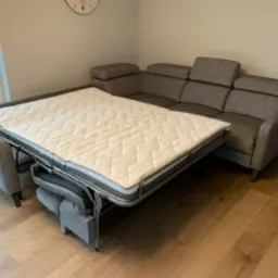 Szara narożna sofa z funkcją spania, rozłożona do pozycji łóżka, widoczny mechanizm rozkładania i materac z pikowaniem, na drewnianej podłodze.