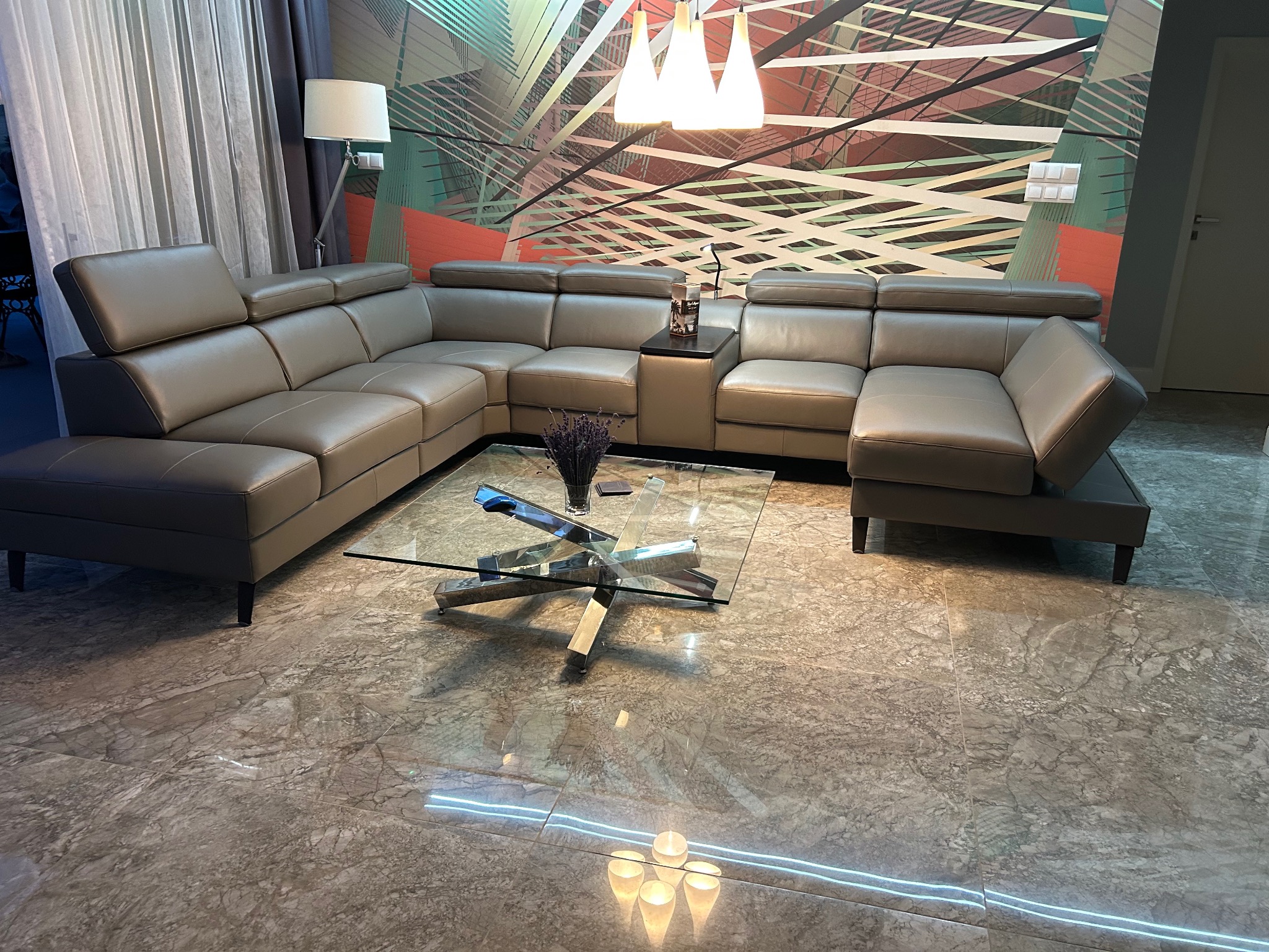 Rozłożysty, skórzany narożnik w odcieniach szarości z regulowanymi zagłówkami, ustawiony w salonie z marmurową podłogą i abstrakcyjną tapetą, oświetlony lampą podłogową i wiszącymi lampami.