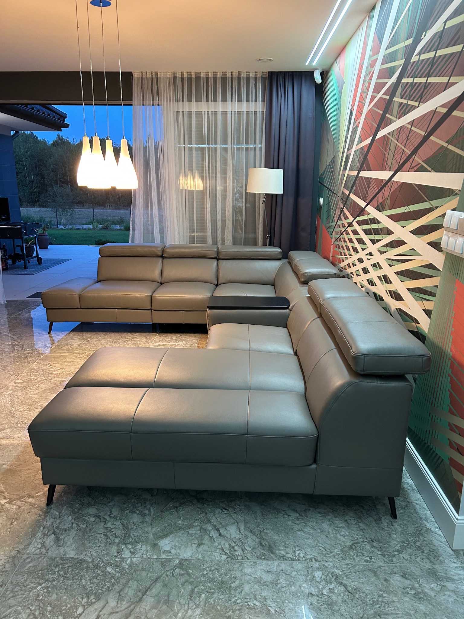 Szary, modułowy narożnik z regulowanymi zagłówkami i czarnym blatem bocznym, w nowoczesnym salonie z marmurową podłogą i abstrakcyjną tapetą.