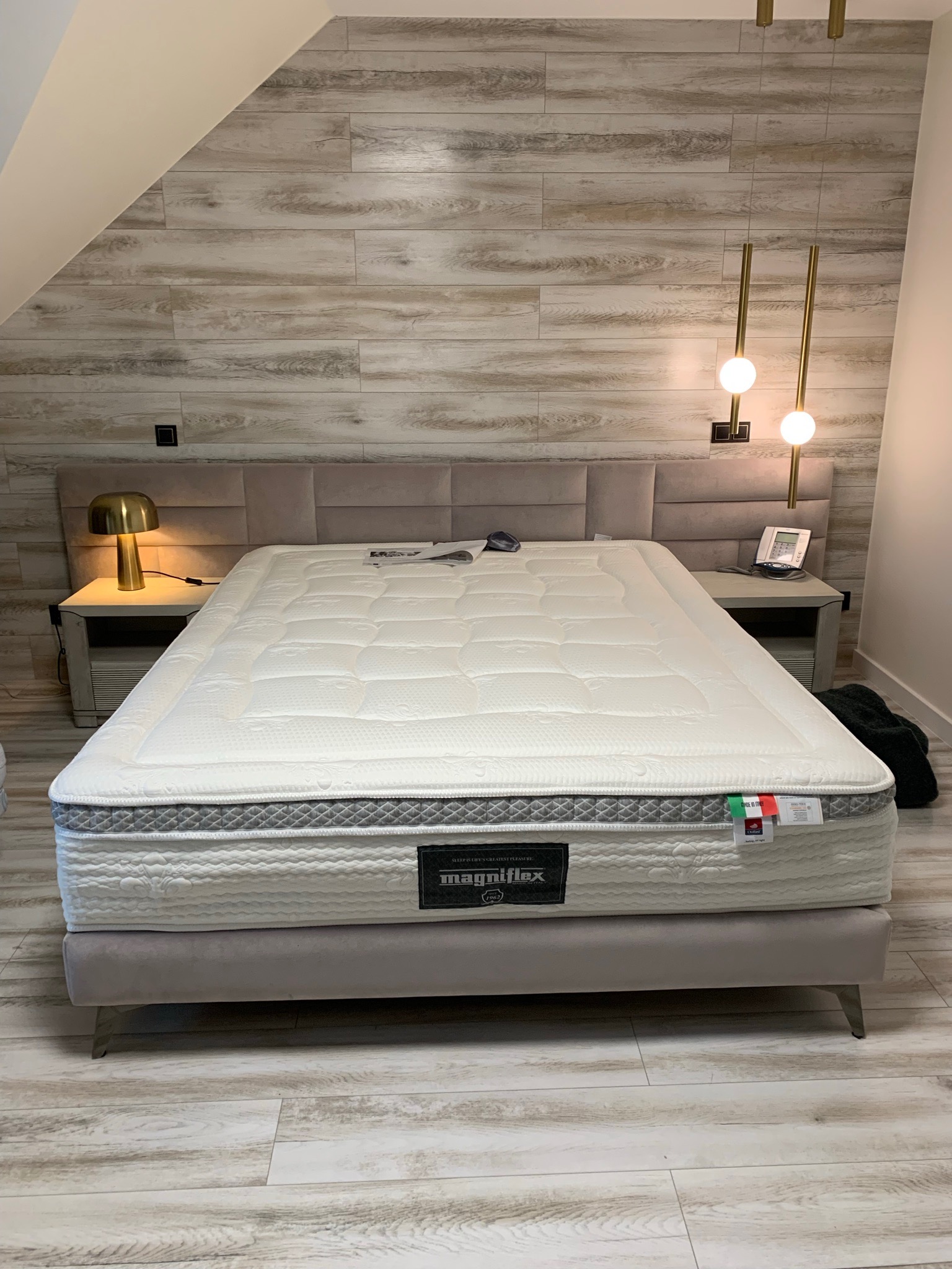 Nowoczesna sypialnia na poddaszu z łóżkiem kontynentalnym Magniflex, beżowym zagłówkiem, drewnopodobną ścianą i złotymi lampami wiszącymi.