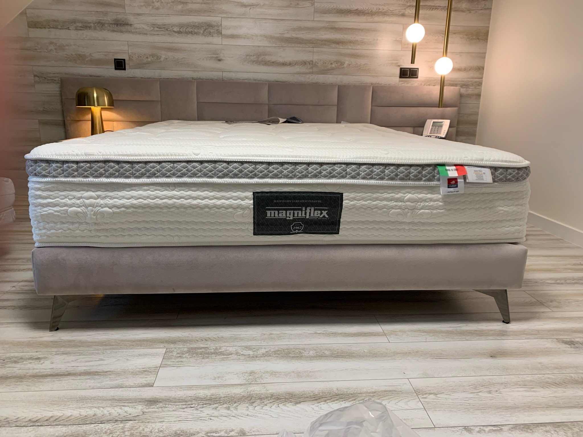 Materac marki Magniflex na łóżku z tapicerowanym wezgłowiem w pokoju z drewnopodobną podłogą i złotą lampą.