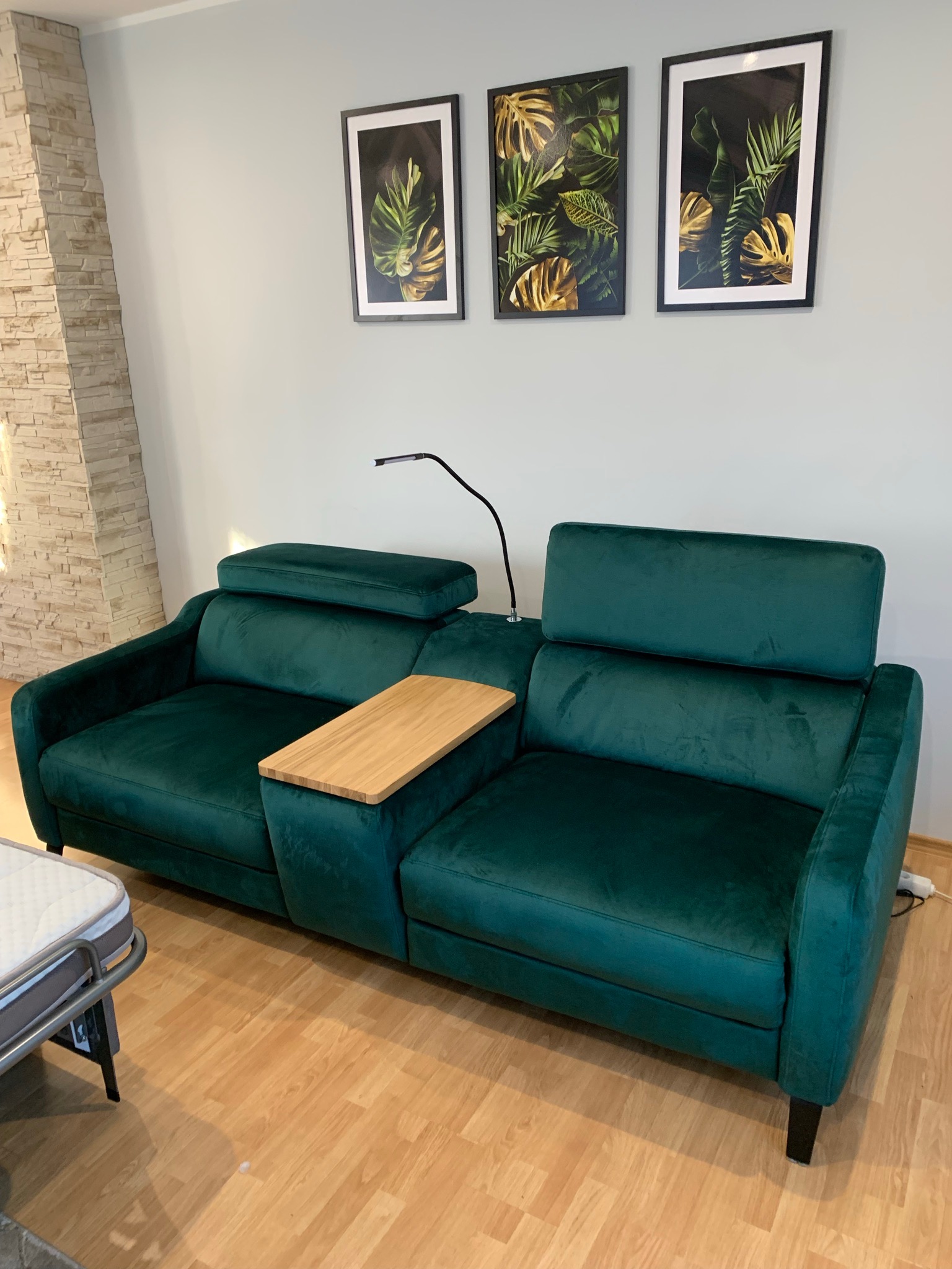 Zielona, welurowa sofa z regulowanymi zagłówkami i drewnianym stolikiem bocznym, ustawiona w salonie z parkietem i obrazami roślinnymi na ścianie.
