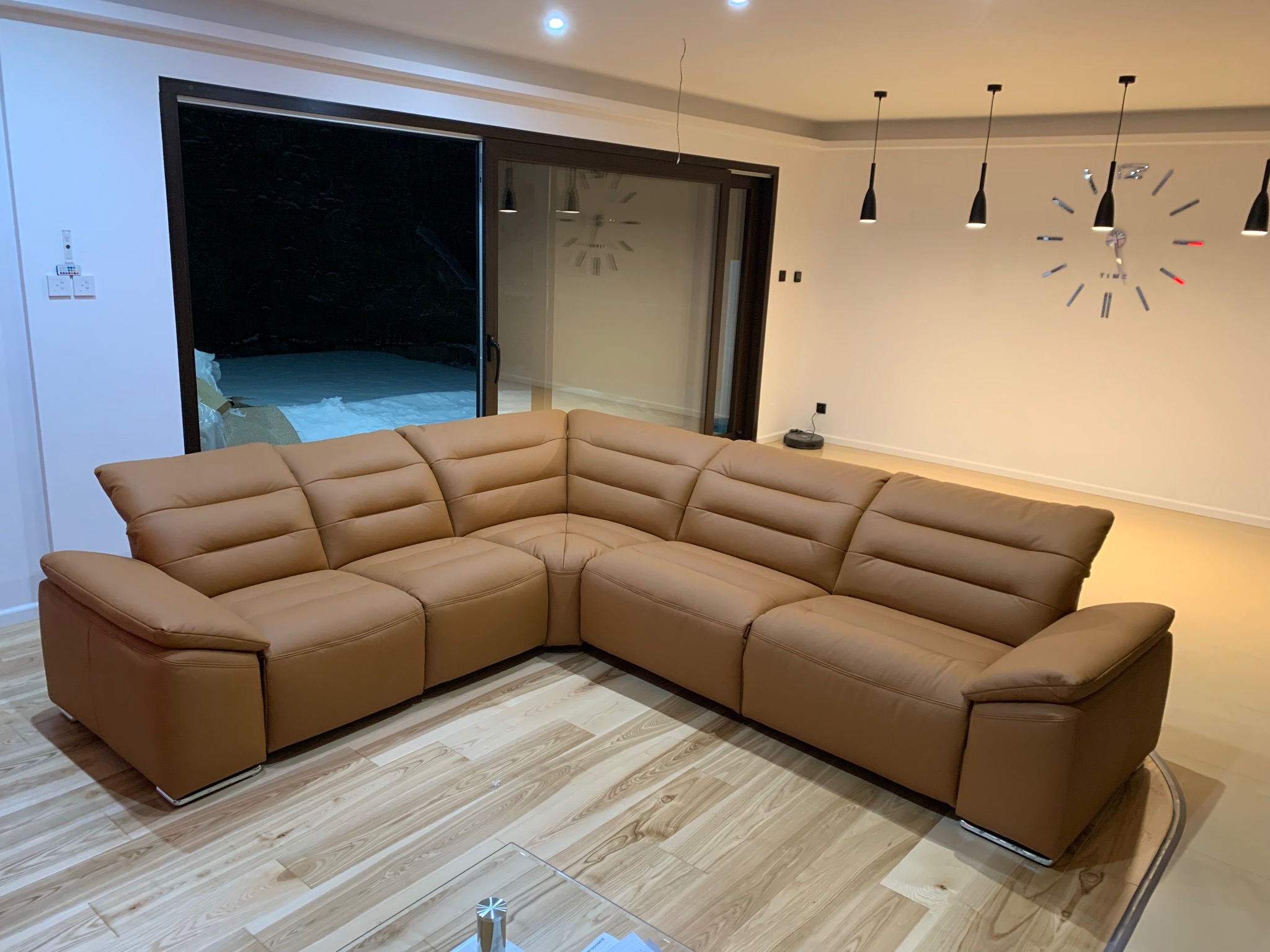 Duży, narożny, skórzany, brązowy wypoczynek z funkcją relaks, stojący w nowoczesnym salonie z drewnianą podłogą i dekoracyjnym zegarem na ścianie.