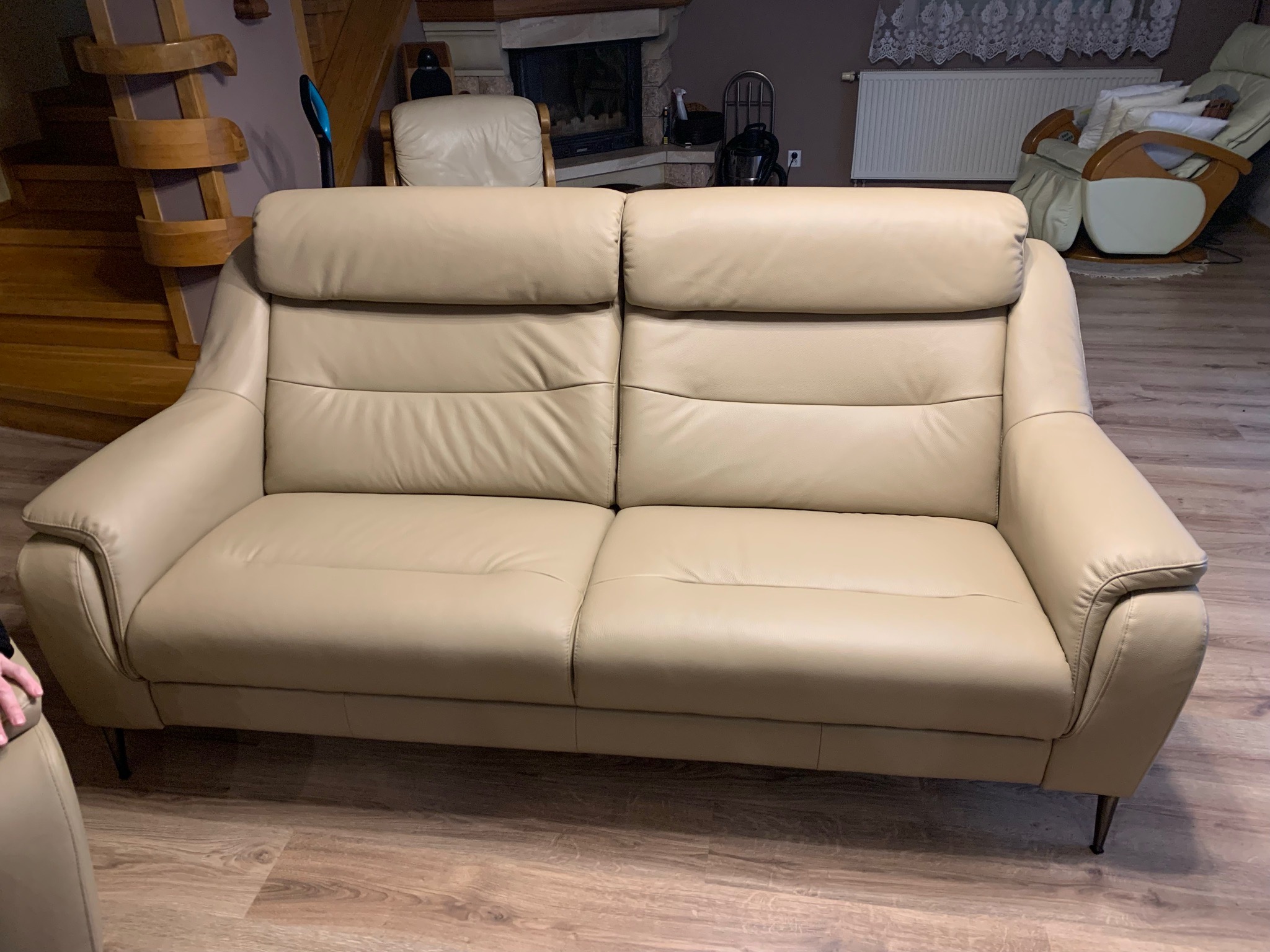 Beżowa, skórzana sofa dwuosobowa z wysokim zagłówkiem, ustawiona na drewnianej podłodze w salonie z kominkiem w tle.