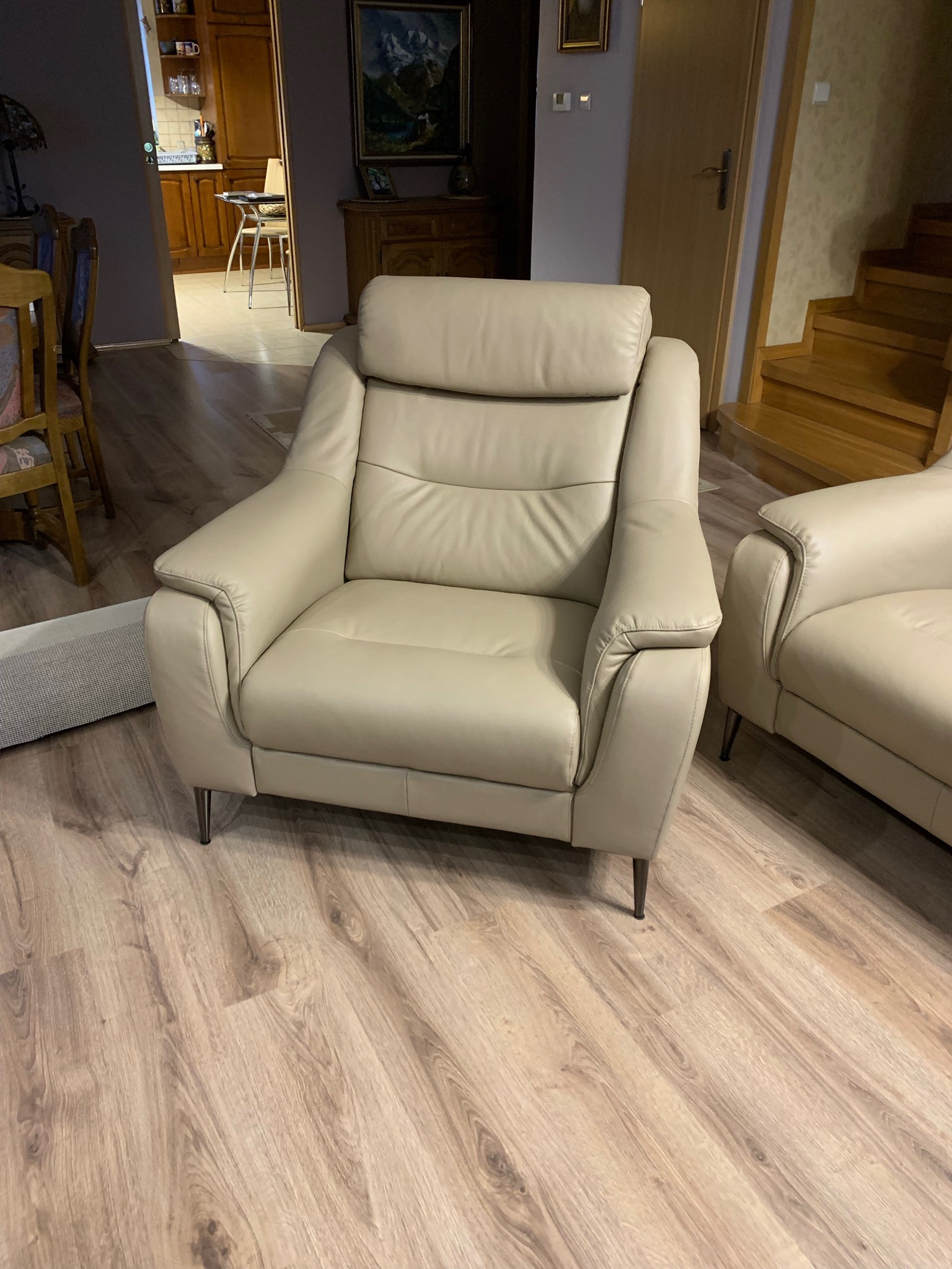 Beżowy, skórzany fotel z wysokim oparciem i metalowymi nóżkami na drewnianej podłodze, widoczne schody i fragment drugiego fotela.