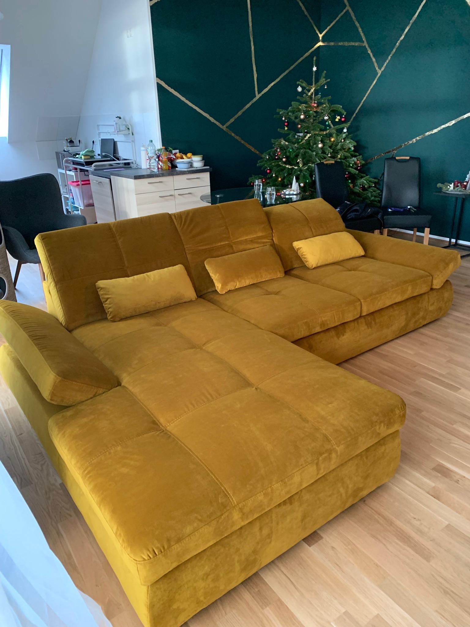 Musztardowa, welurowa sofa narożna z poduszkami, na tle zielonej ściany z geometrycznym wzorem i choinki.