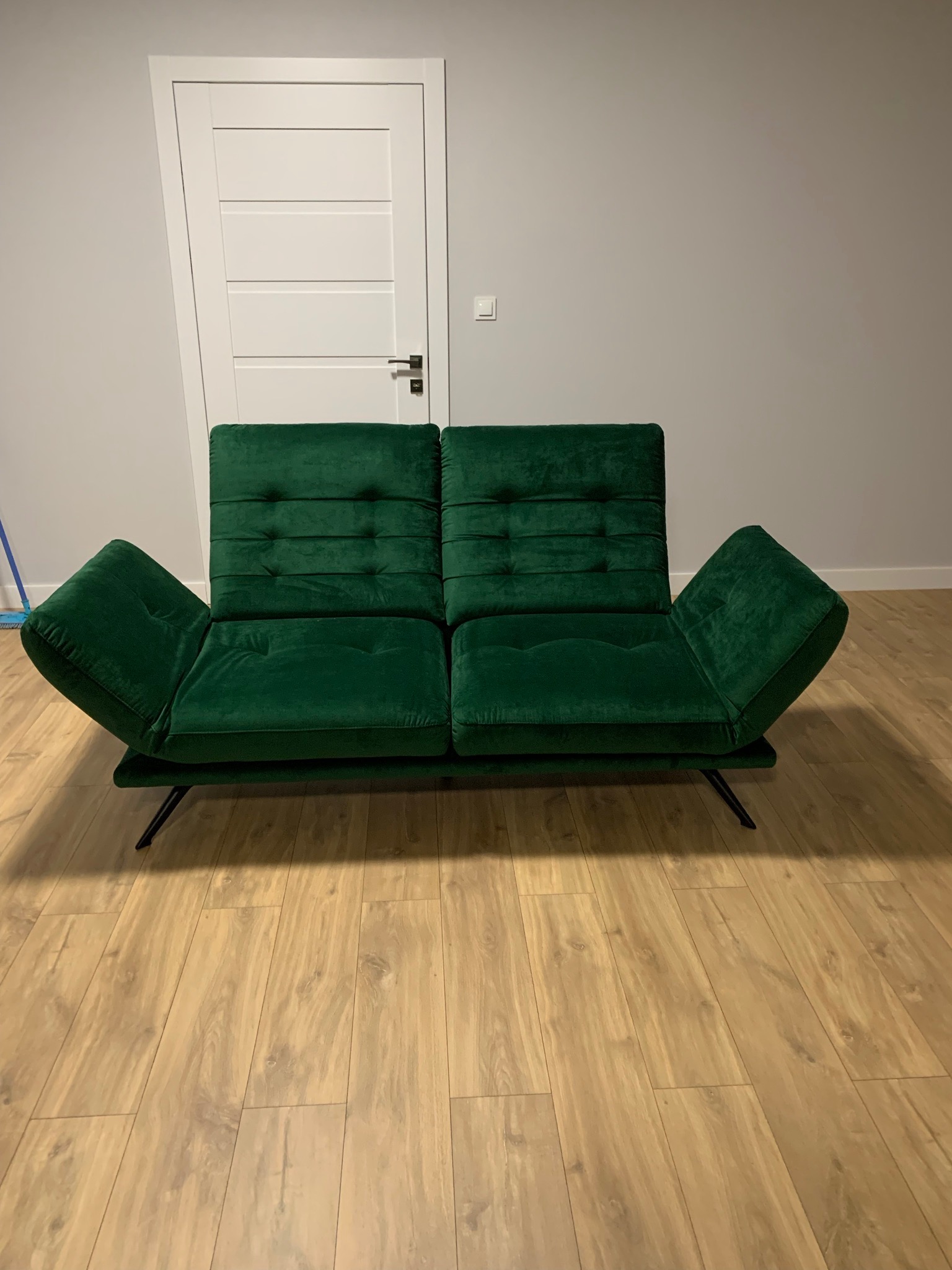 Rozkładana sofa w kolorze butelkowej zieleni, z pikowanymi oparciami i podłokietnikami, na cienkich czarnych nóżkach, ustawiona na panelach podłogowych, z białą ścianą i drzwiami w tle.