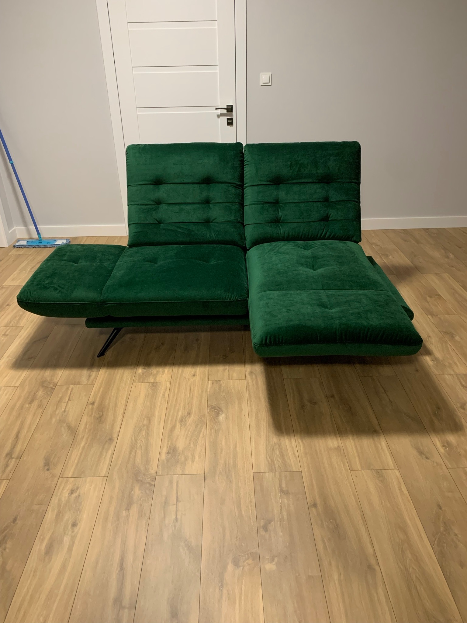 Rozłożona, dwuosobowa sofa tapicerowana aksamitną tkaniną w kolorze butelkowej zieleni, z pikowanymi oparciami i siedziskami, ustawiona na panelach podłogowych, widoczny fragment mopa oparty o ścianę.