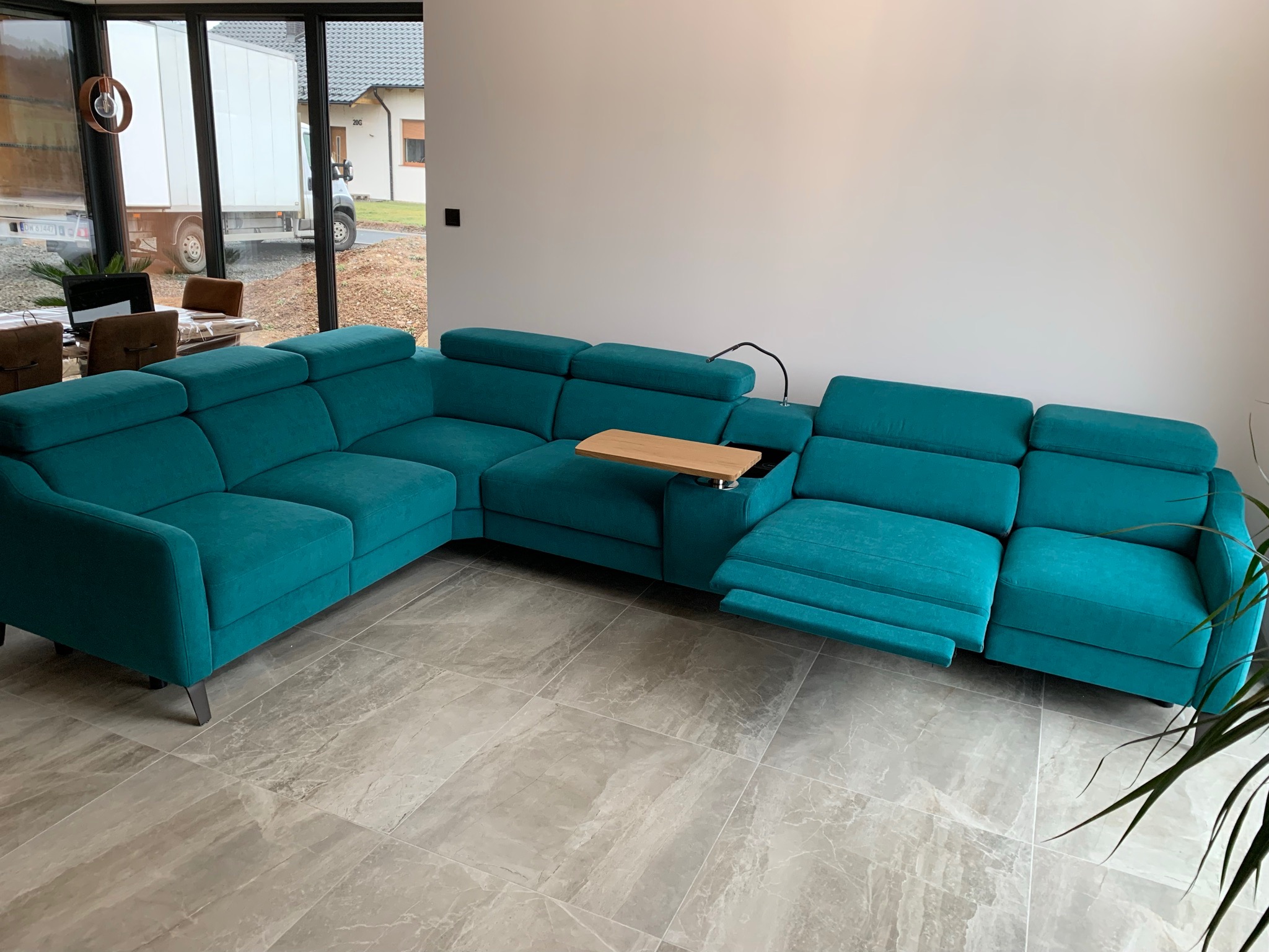 Turkusowy narożnik z funkcją relaks, wysuwanym podnóżkiem i drewnianym blatem, ustawiony na marmurowej podłodze w nowoczesnym salonie z widokiem na zaparkowany samochód dostawczy.