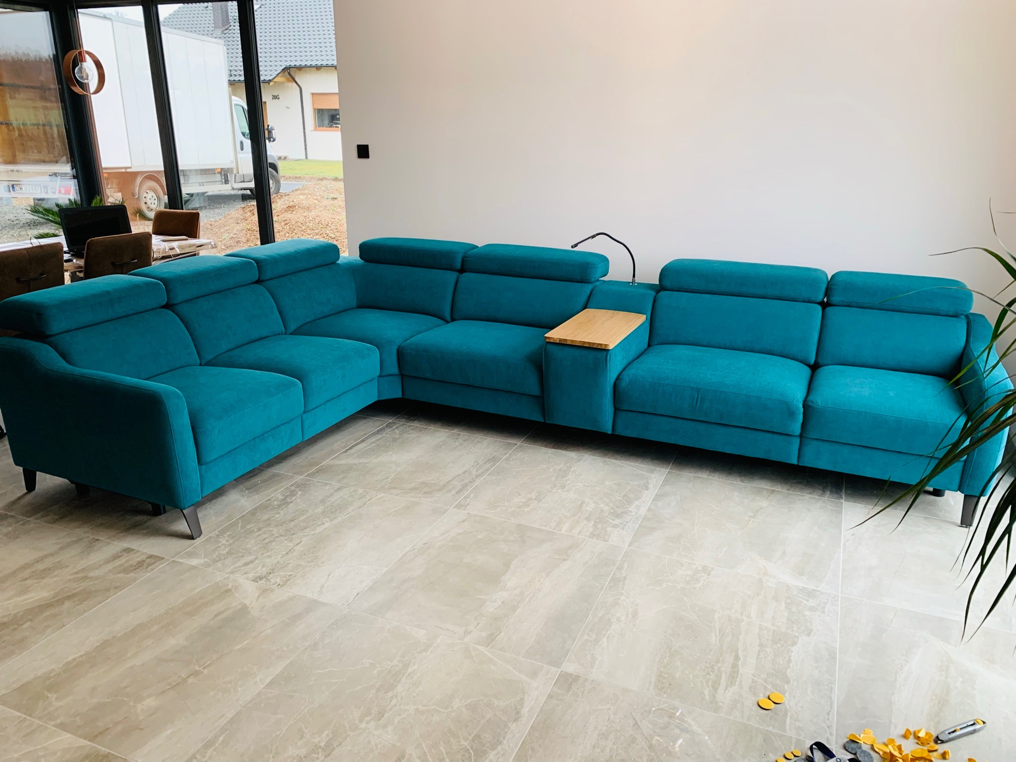 Turkusowy, narożny zestaw wypoczynkowy z drewnianym panelem bocznym, ustawiony na marmurowej podłodze w przestronnym salonie z widokiem na samochód dostawczy za oknem.