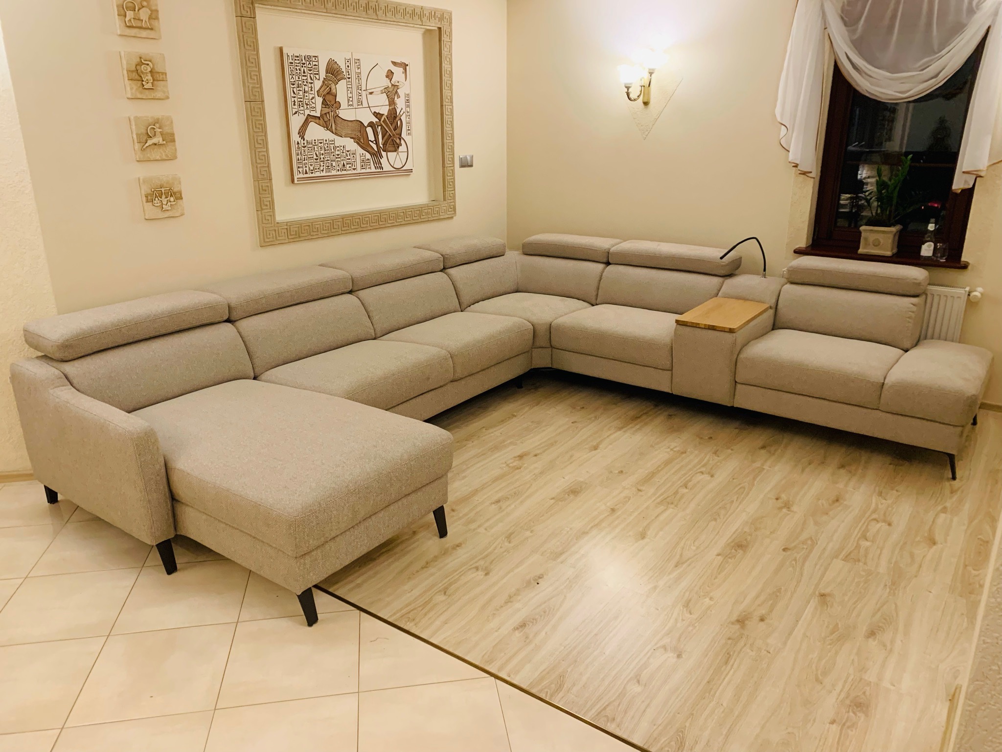 Jasnoszary narożnik z regulowanymi zagłówkami, drewnianym stolikiem bocznym i czarnymi nóżkami, w salonie z parkietem i dekoracyjnymi obrazami.