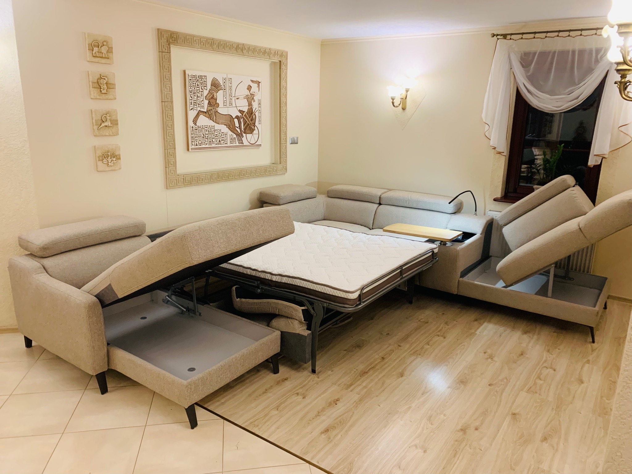 Rozłożona narożna sofa z beżową tapicerką, widoczne pojemniki na pościel, materac na metalowej ramie, w tle ściana z obrazami w stylu egipskim.