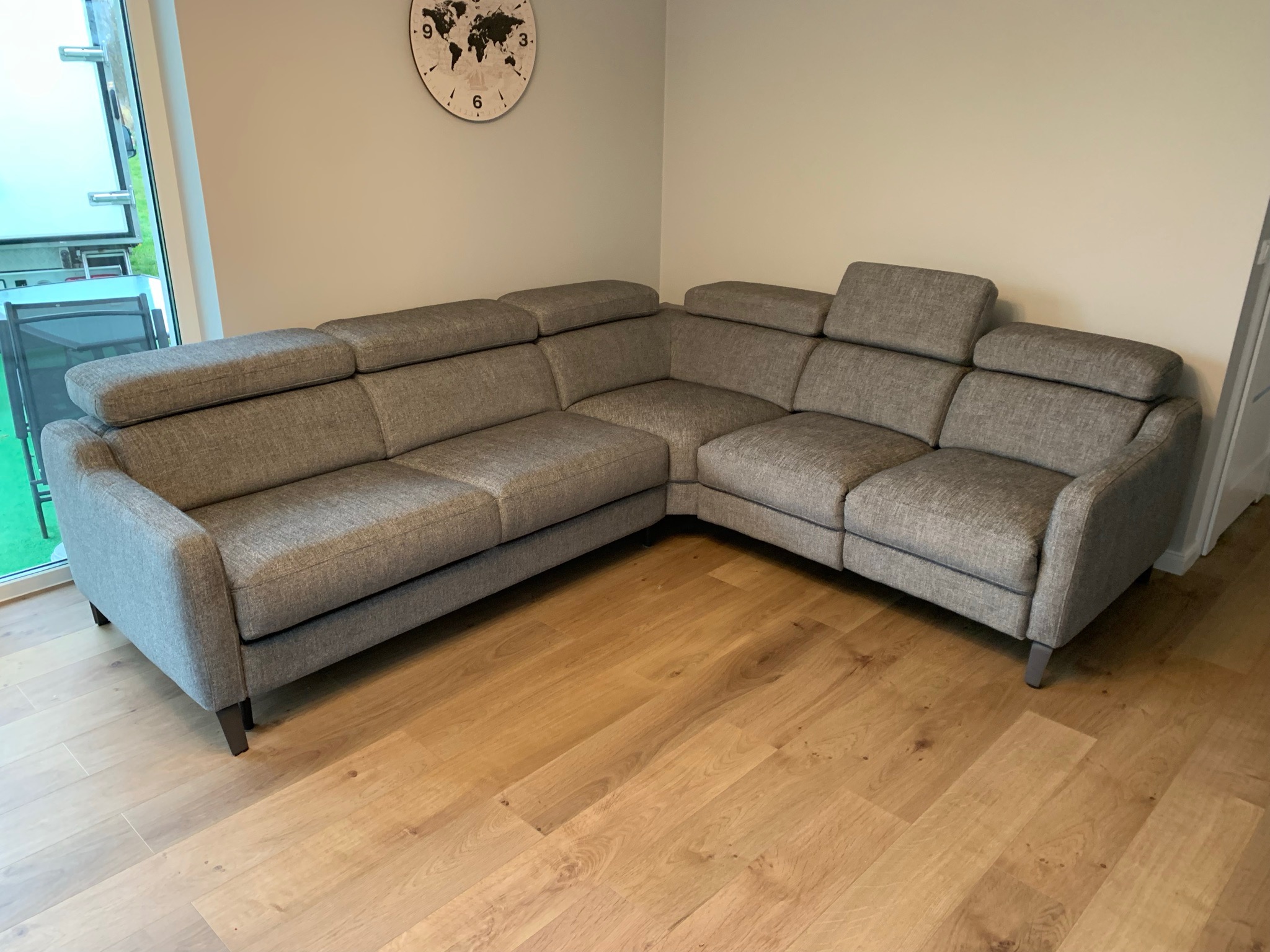 Szara narożna sofa z regulowanymi zagłówkami na drewnianej podłodze, widoczny zegar z mapą świata na ścianie.