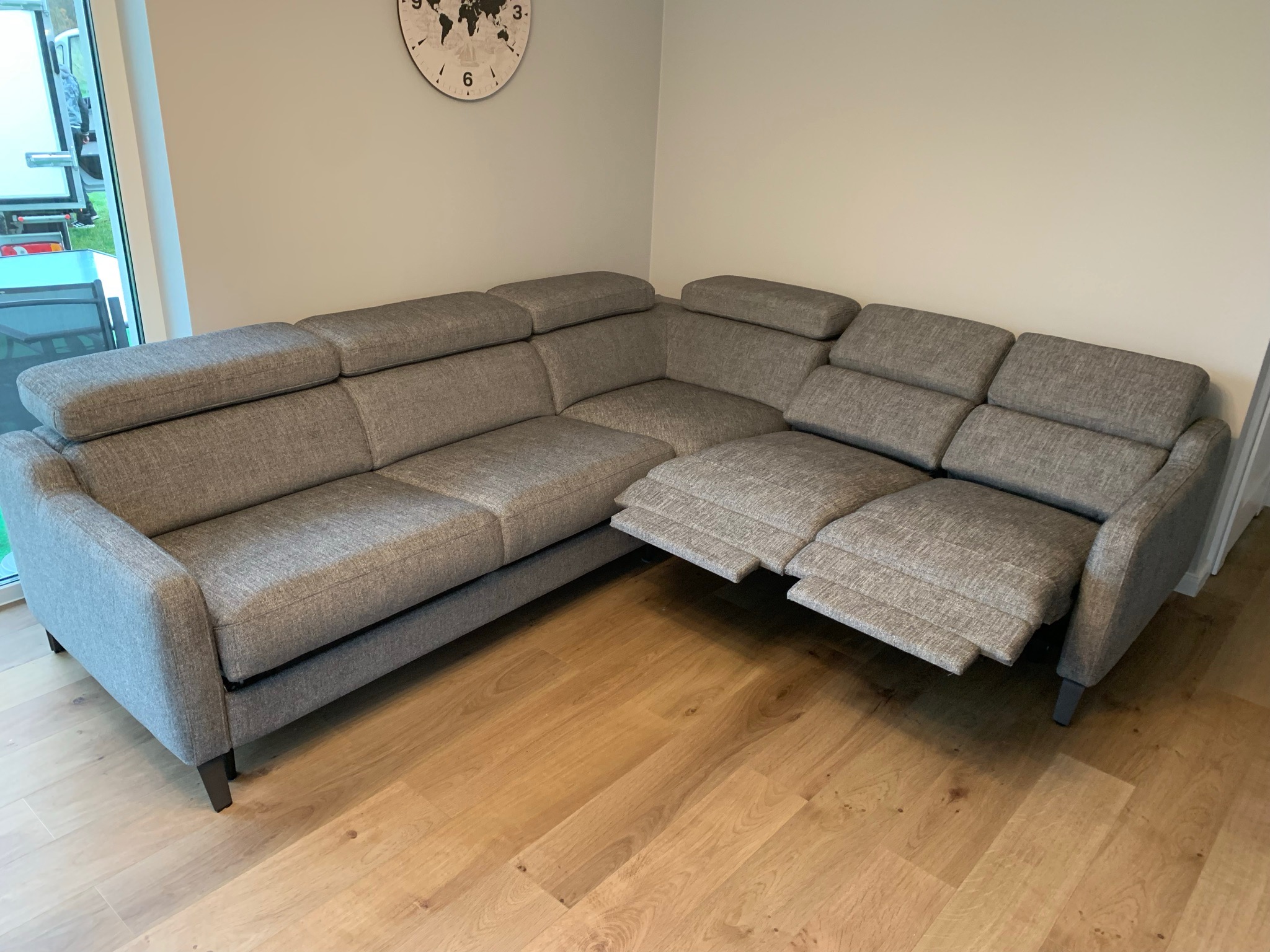 Szara, narożna sofa tapicerowana z regulowanymi zagłówkami i wysuwanym podnóżkiem, ustawiona na drewnianej podłodze w jasnym wnętrzu, widok z boku.