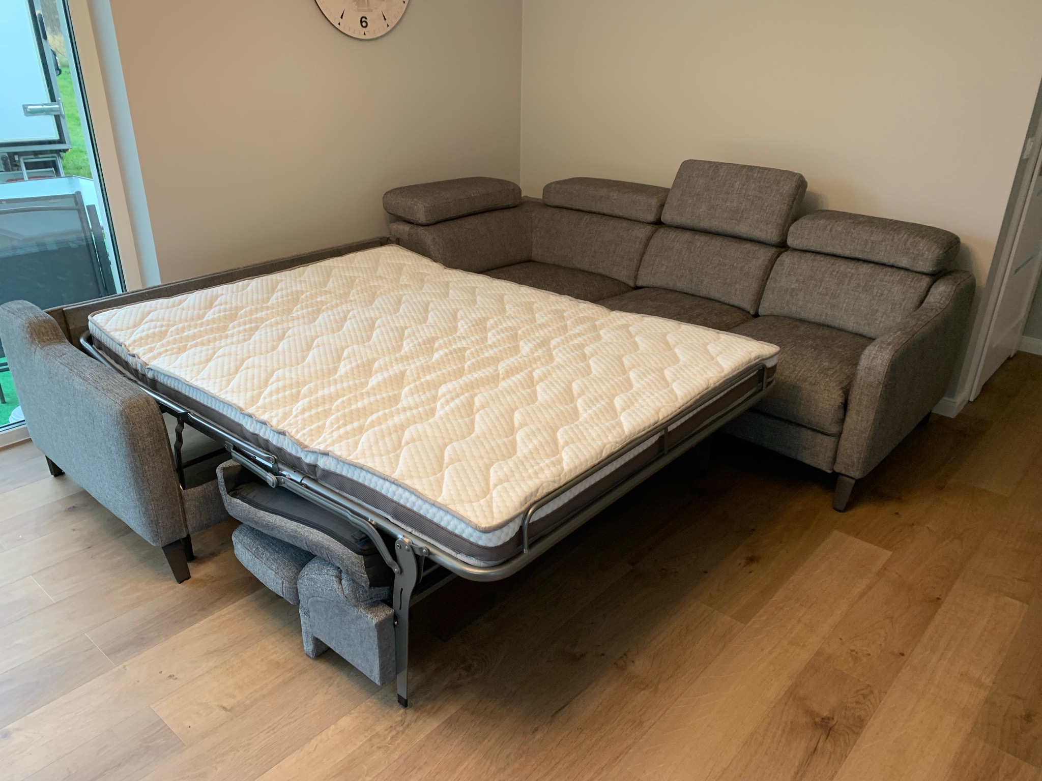 Szara narożna sofa z funkcją spania, rozłożona do pozycji łóżka, widoczny mechanizm rozkładania i materac z pikowaniem, na drewnianej podłodze.