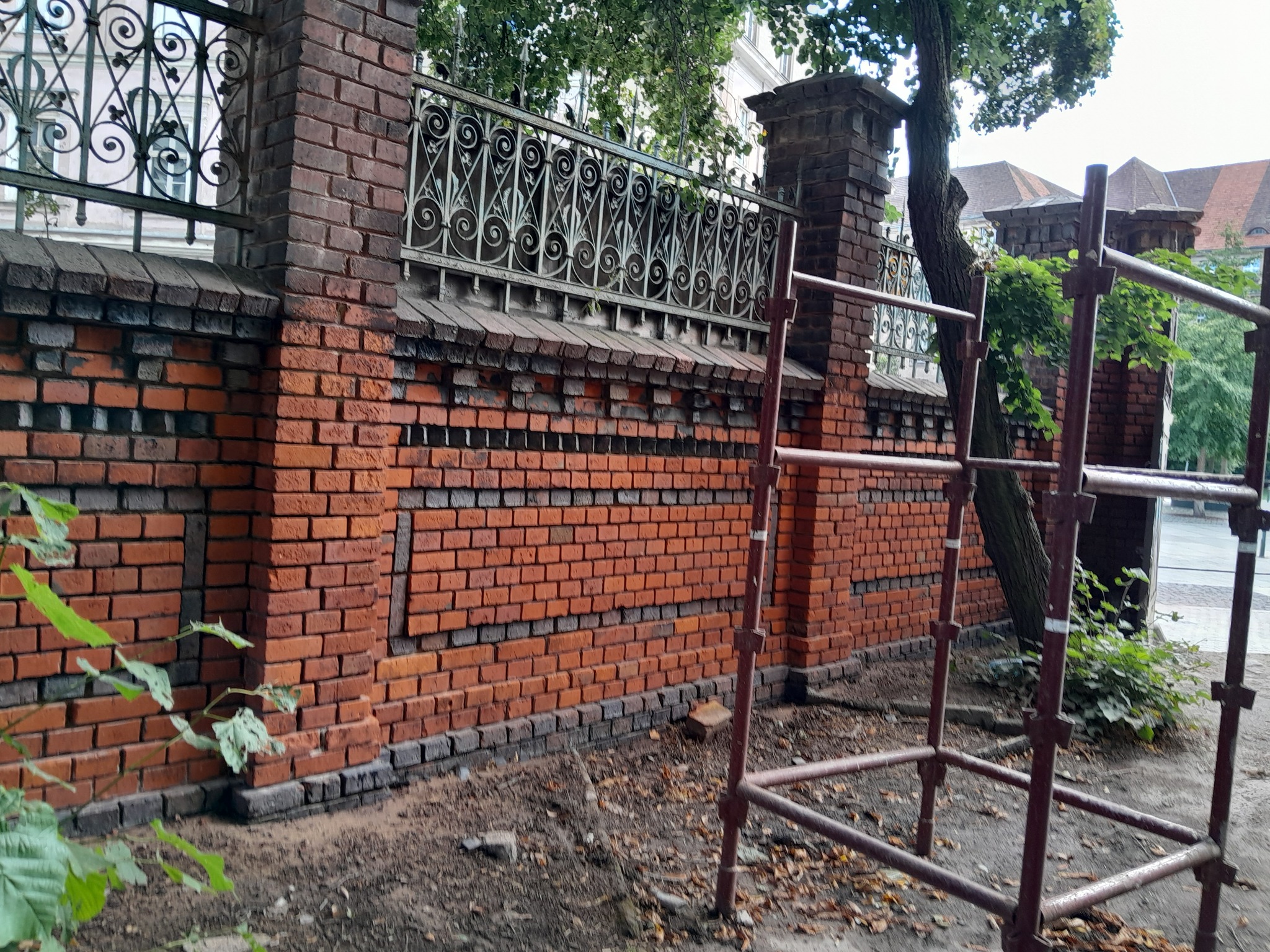 Fragment ceglanego muru z ozdobną metalową kratą, częściowo odnowiony, z widocznymi rusztowaniami.