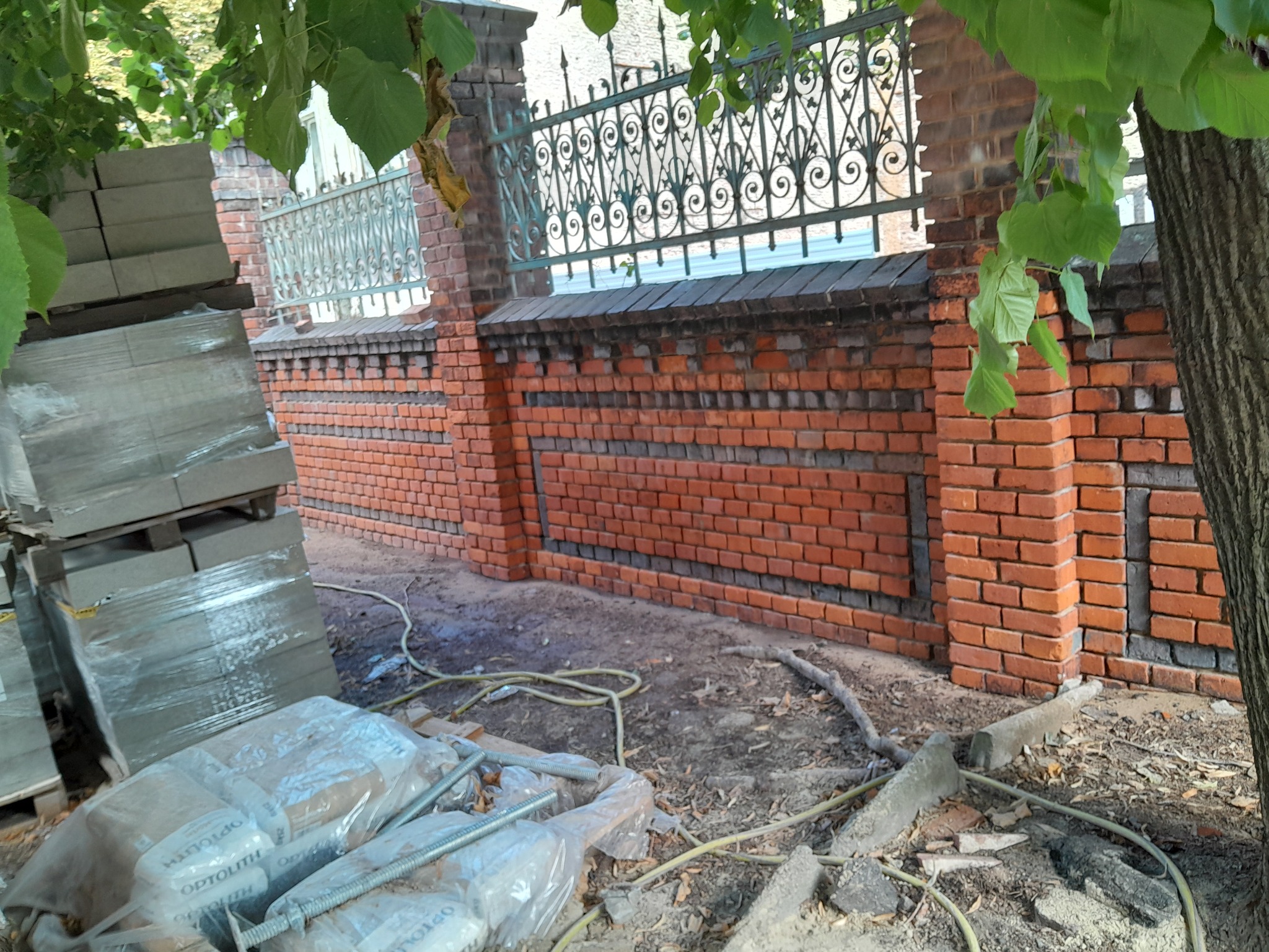 Wznoszenie fragmentu muru z czerwonej cegły z dekoracyjnym, metalowym ogrodzeniem w tle, widoczne materiały budowlane i narzędzia.