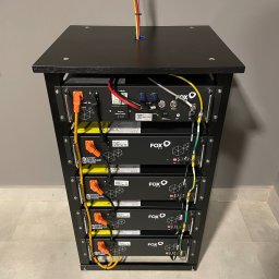 Sollogy - Czarny regał z modułami magazynu energii FOX ESS HV2600, widoczne okablowanie i złącza, tło szara ściana i jasna podłoga.