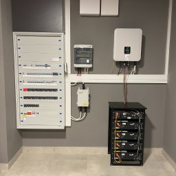 Sollogy - Kompletna instalacja z magazynem energii: rozdzielnica elektryczna, licznik, inwerter i moduł baterii w czarnej obudowie, zamontowane na szarej ścianie w pomieszczeniu z jasnymi płytkami podłogowymi.