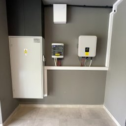 Sollogy - Wewnętrzna instalacja: rozdzielnica elektryczna Nestino, licznik energii i inwerter FoxESS zamontowane na szarej ścianie w pomieszczeniu z szarą podłogą z płytek. Widoczne przewody i rurki...