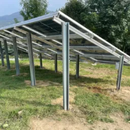 Instalacja paneli słonecznych na metalowej konstrukcji w Krze, widok od dołu z perspektywy trawnika, góry i drzewa w tle.