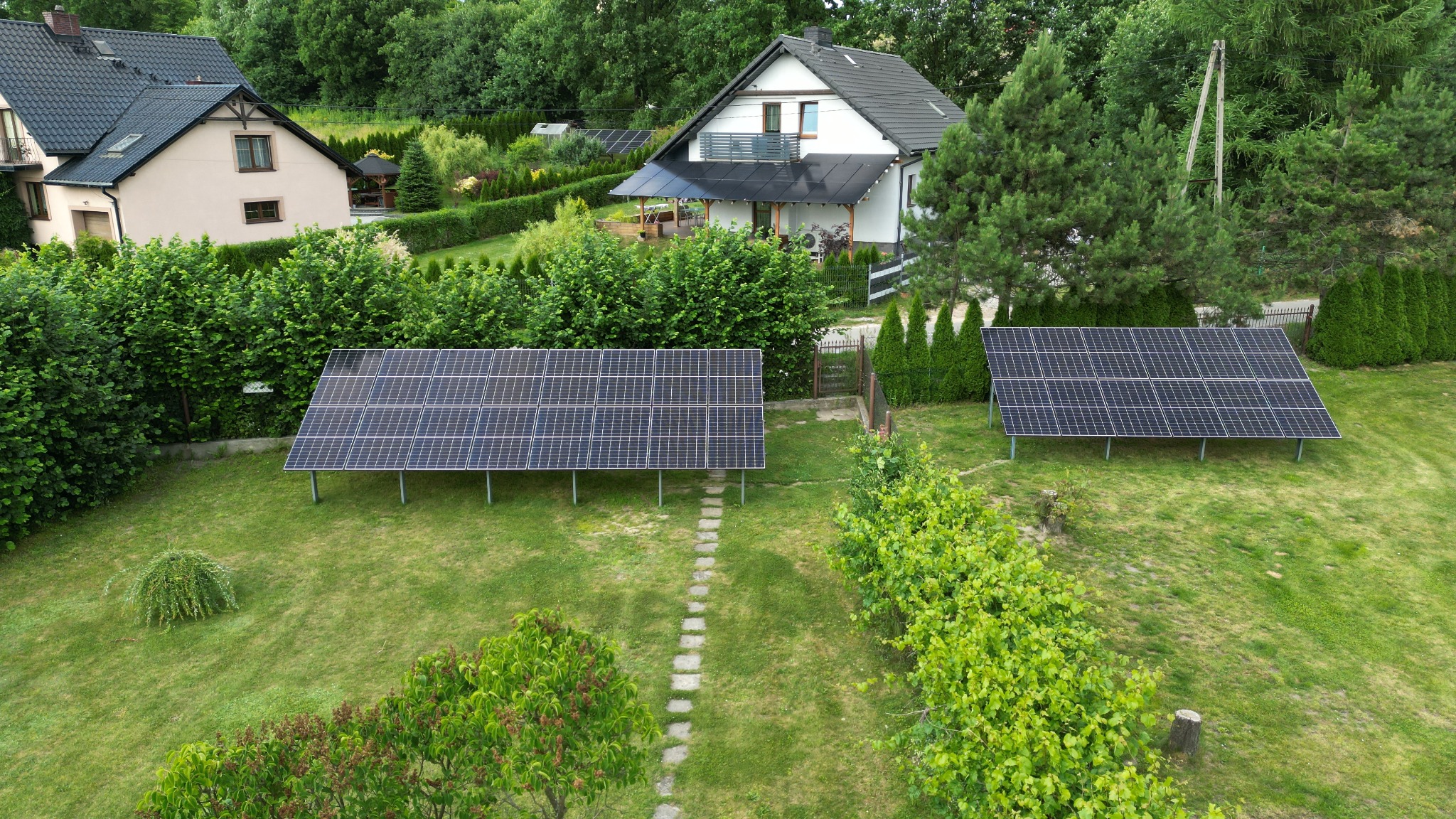 Dwa rzędy paneli słonecznych na zielonym trawniku przed domem z panelami na dachu, widok z góry.