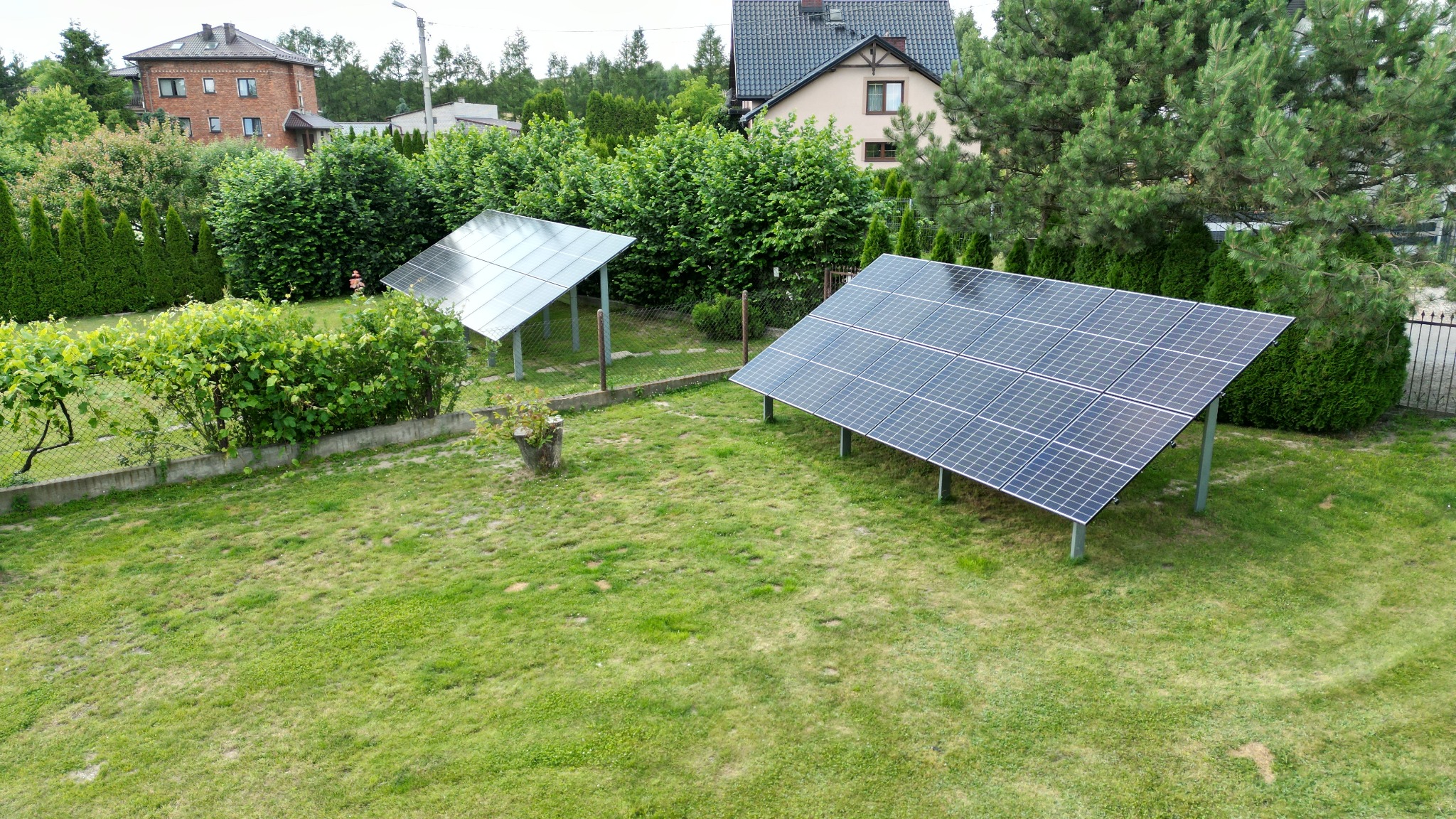 Dwa panele słoneczne na metalowych stelażach, ustawione na trawniku w ogrodzie, widok z góry z perspektywy drona, w tle domy jednorodzinne i drzewa.