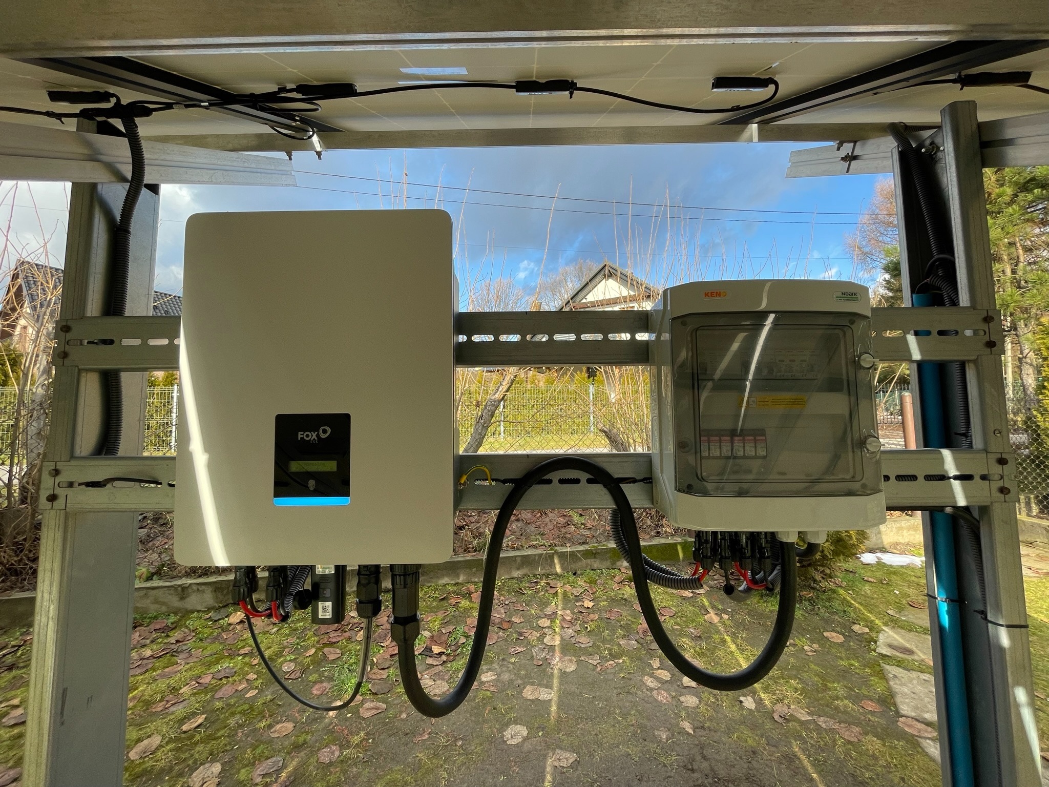 Instalacja fotowoltaiczna: inwerter FoxESS z podświetlonym na niebiesko ekranem i skrzynka zabezpieczeń KENO zamontowane na metalowej konstrukcji na tle częściowo widocznego nieba i zabudowań.