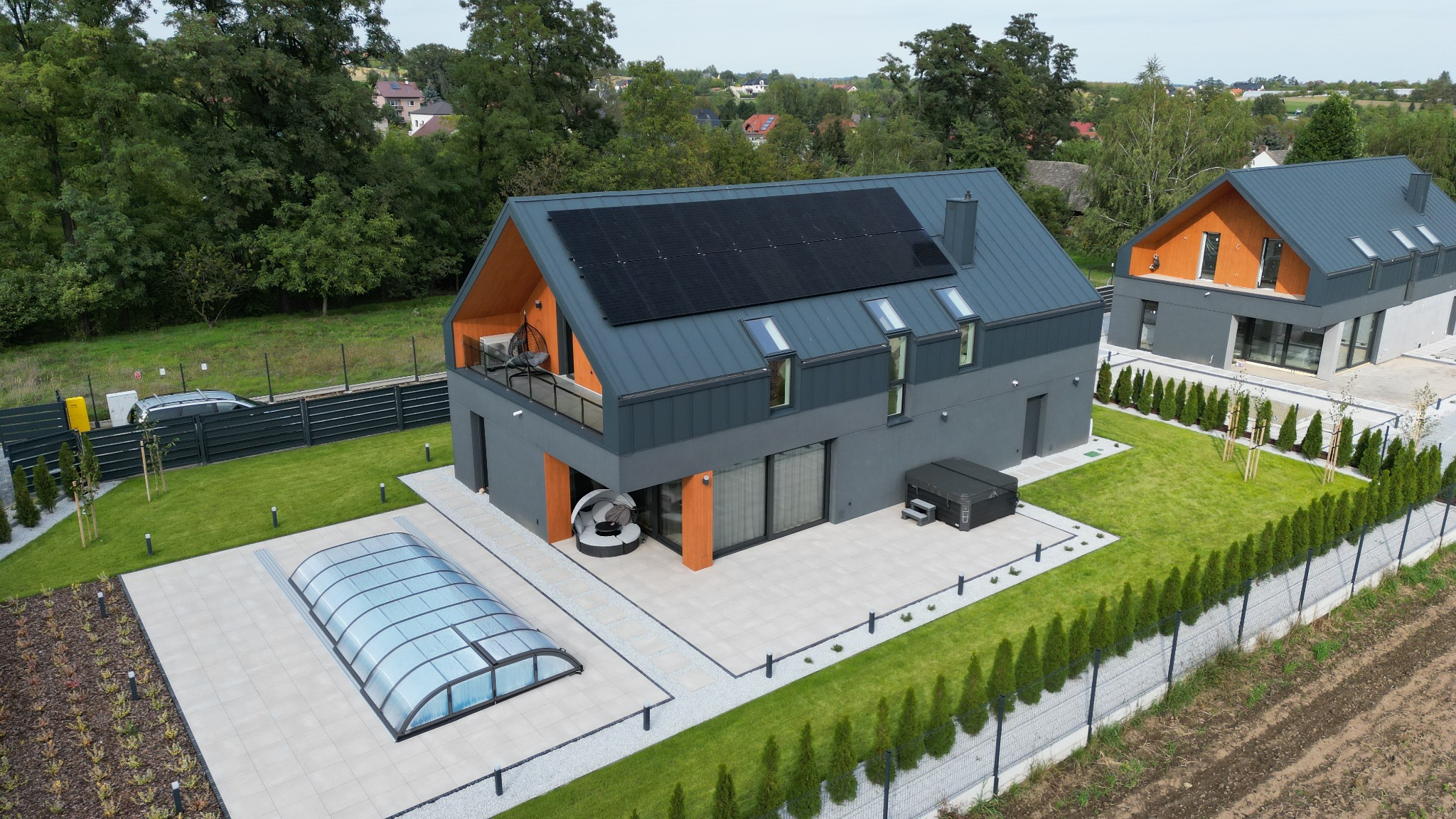 Nowoczesny dom z szarą elewacją i panelami słonecznymi na dachu, widok z lotu ptaka, zadaszony basen obok domu, zadbany trawnik i tuje wzdłuż ogrodzenia.