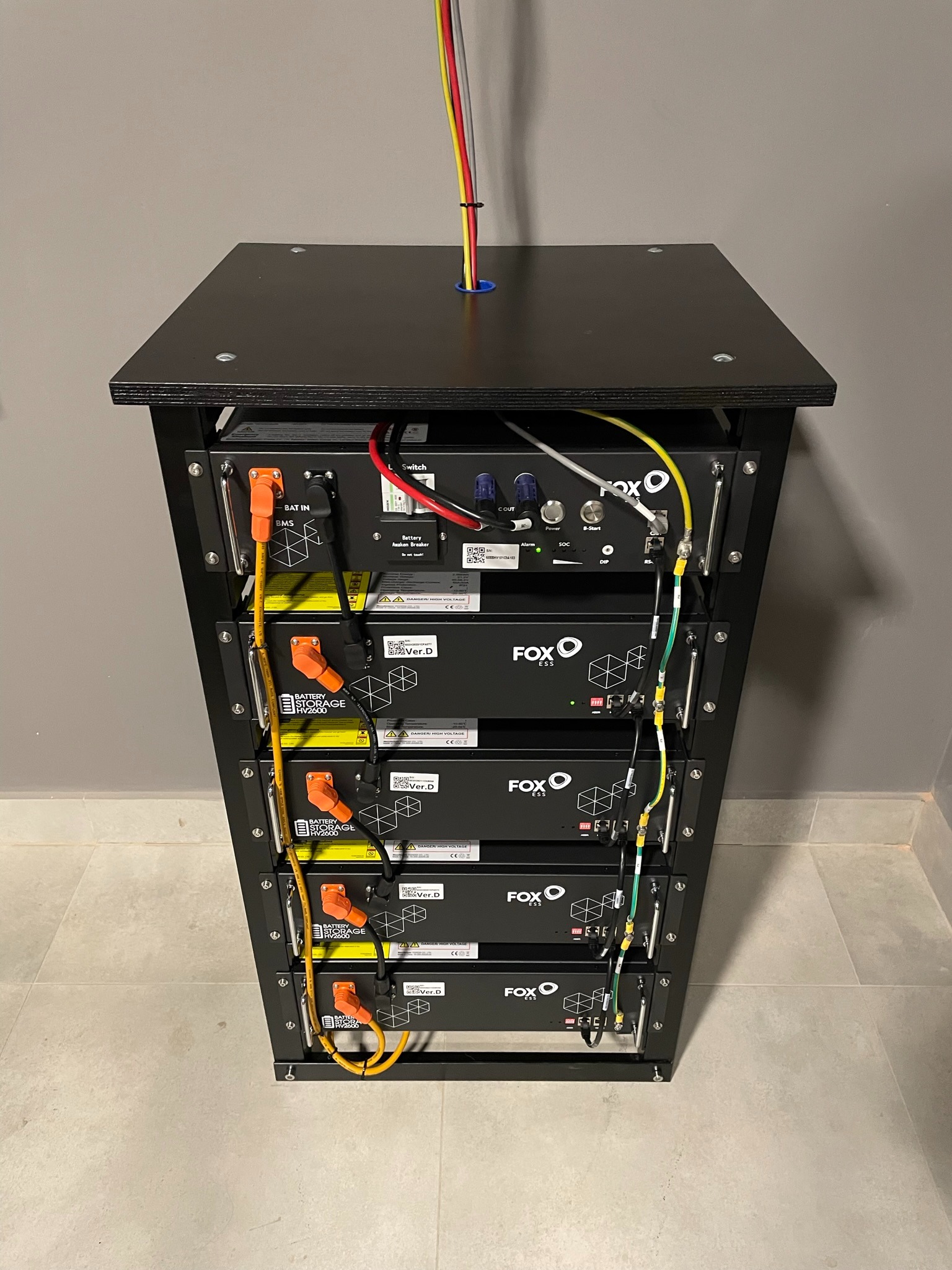 Czarny regał z modułami magazynu energii FOX ESS HV2600, widoczne okablowanie i złącza, tło szara ściana i jasna podłoga.