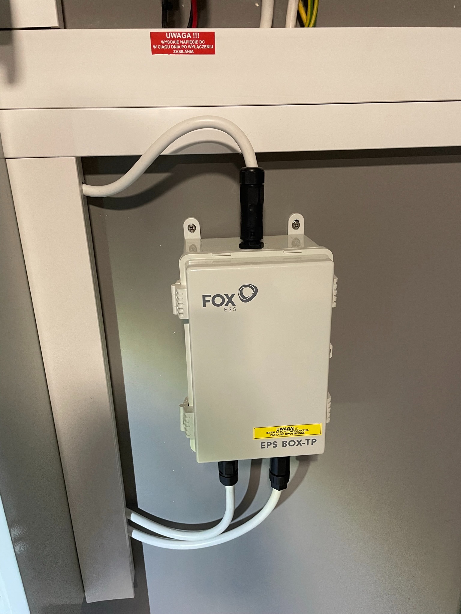 Biała skrzynka przyłączeniowa EPS BOX-TP firmy FOX ESS, zamontowana na szarej ścianie, z podłączonymi białymi kablami. Widoczna naklejka ostrzegawcza o instalacji fotowoltaicznej i zasilaniu...