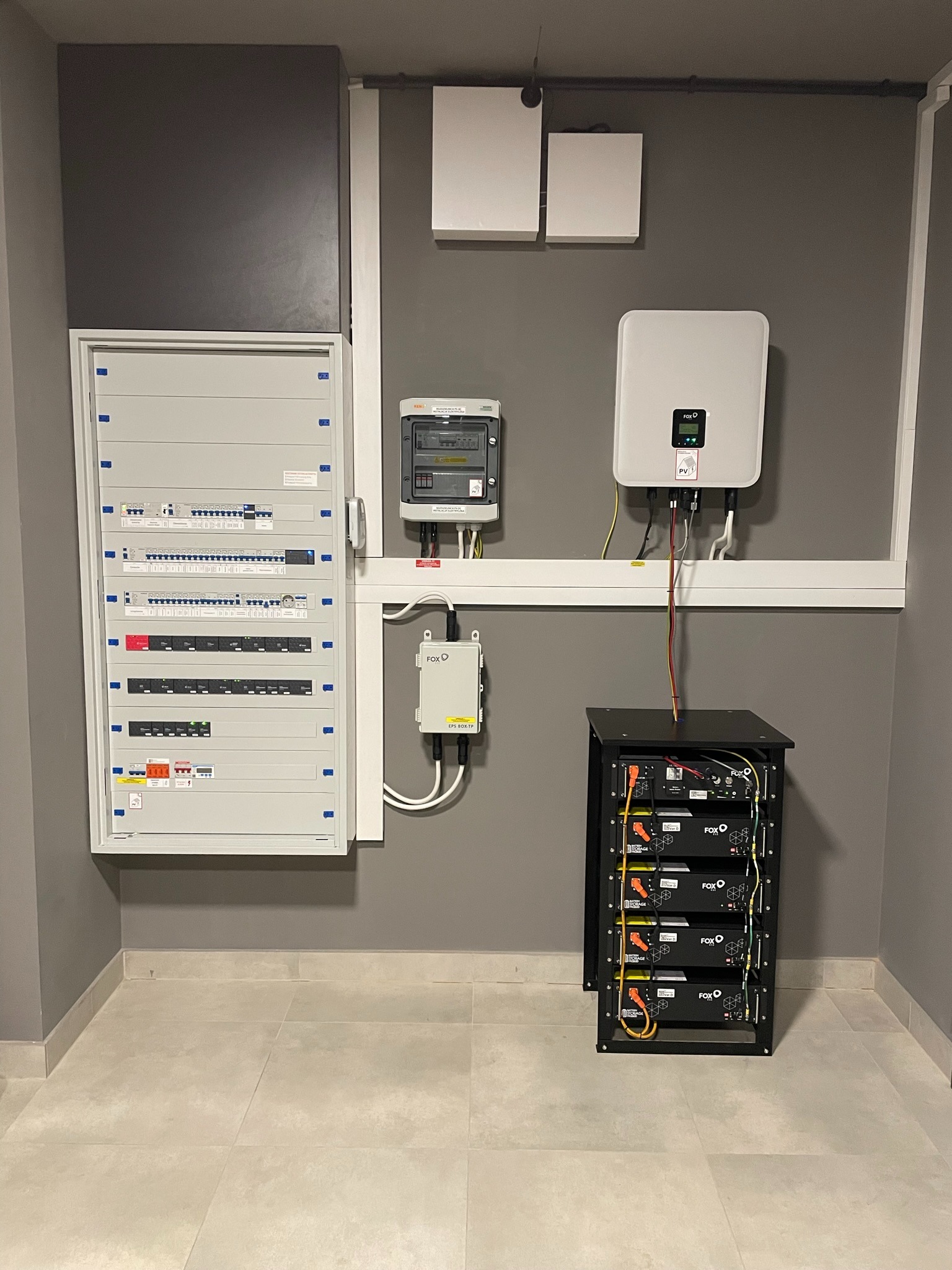 Kompletna instalacja z magazynem energii: rozdzielnica elektryczna, licznik, inwerter i moduł baterii w czarnej obudowie, zamontowane na szarej ścianie w pomieszczeniu z jasnymi płytkami podłogowymi.