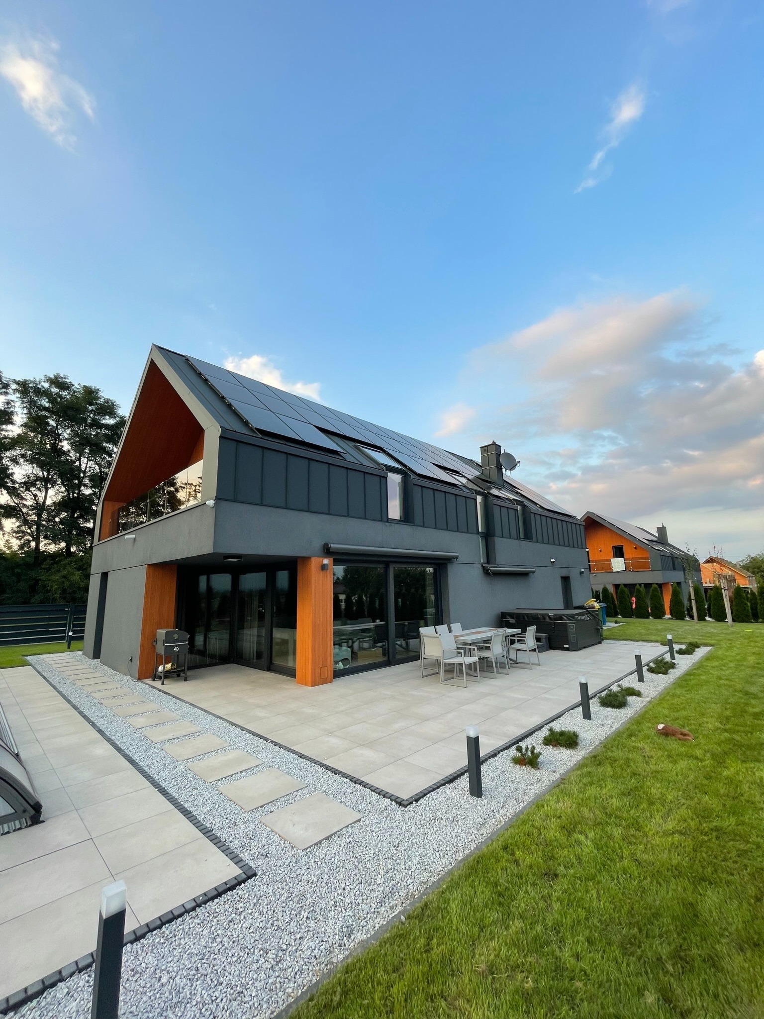 Nowoczesny dom jednorodzinny z panelami słonecznymi na dachu, szara elewacja z drewnianymi akcentami, duży taras z meblami ogrodowymi, zadbany trawnik i oświetlenie zewnętrzne.