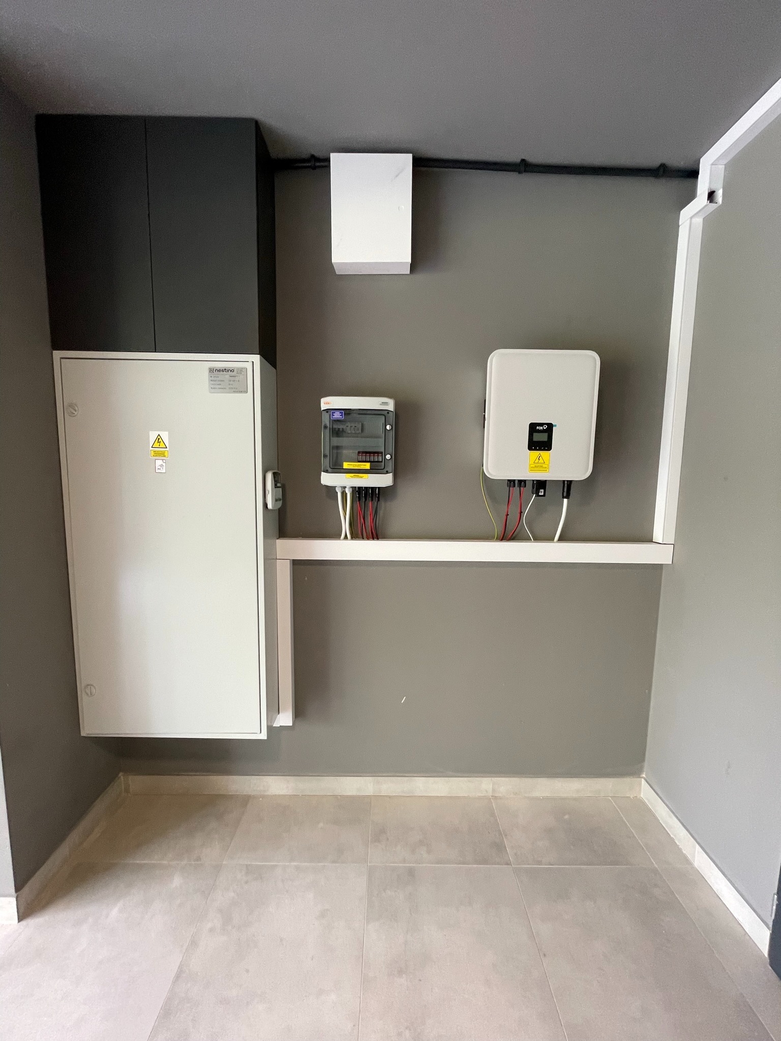 Wewnętrzna instalacja: rozdzielnica elektryczna Nestino, licznik energii i inwerter FoxESS zamontowane na szarej ścianie w pomieszczeniu z szarą podłogą z płytek. Widoczne przewody i rurki...