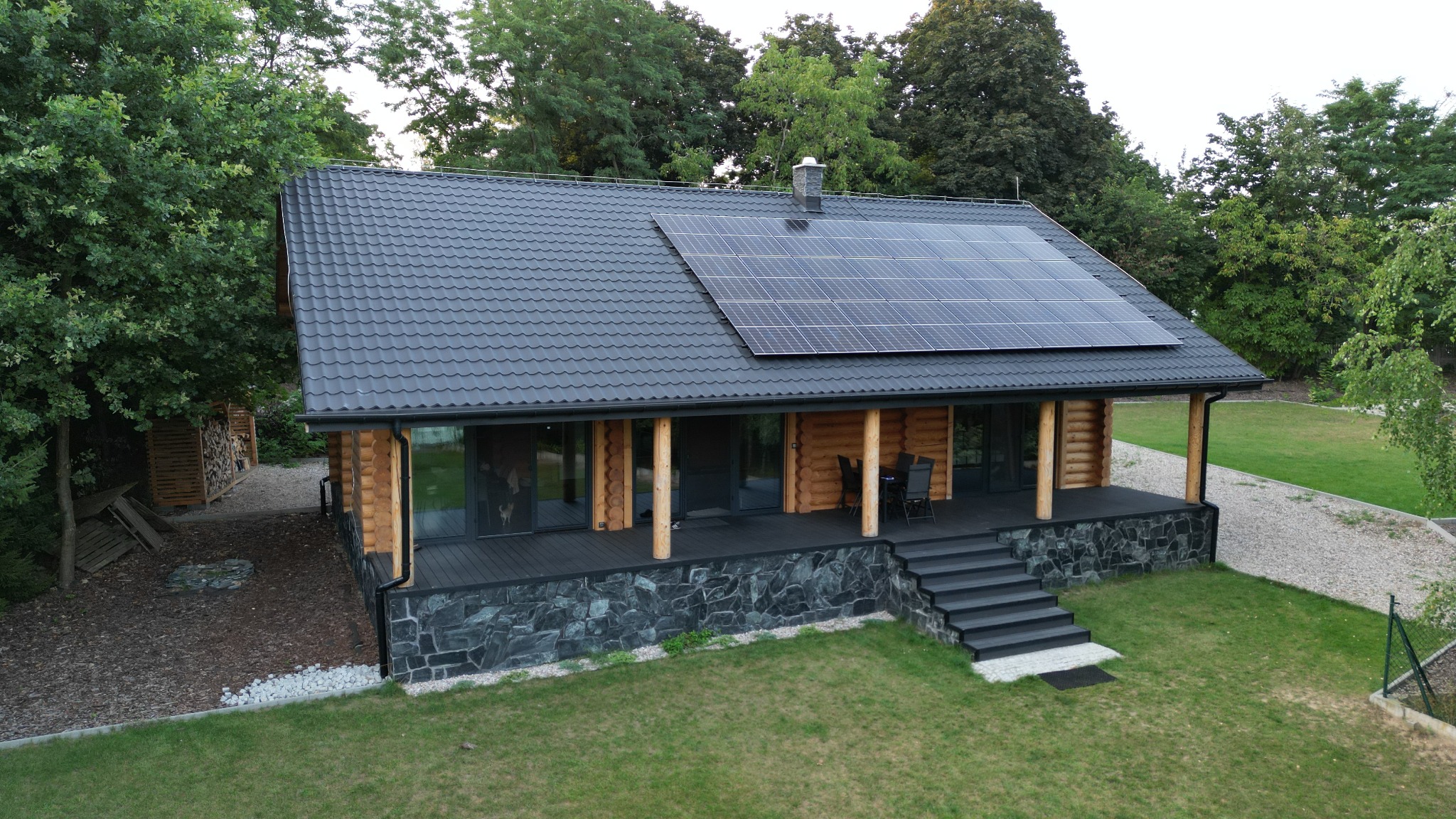 Drewniany dom z ciemną dachówką i panelami fotowoltaicznymi na dachu, otoczony zielenią, widok z lotu ptaka.