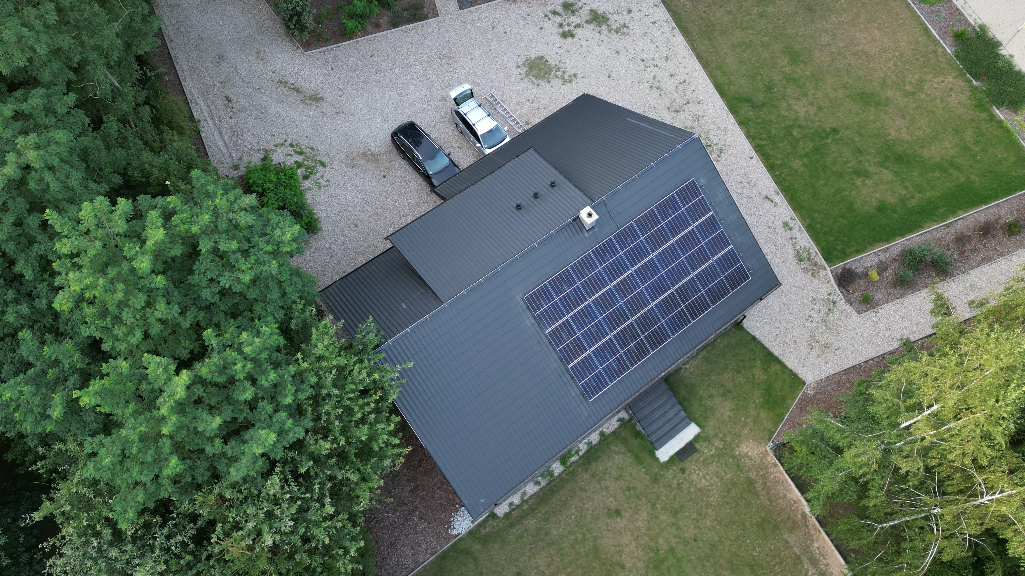 Widok z lotu ptaka na dom z ciemnym dachem i panelami słonecznymi, obok zaparkowane dwa samochody, w tym jeden z otwartym bagażnikiem i drabiną na dachu, otoczony zielenią i żwirowym podjazdem.