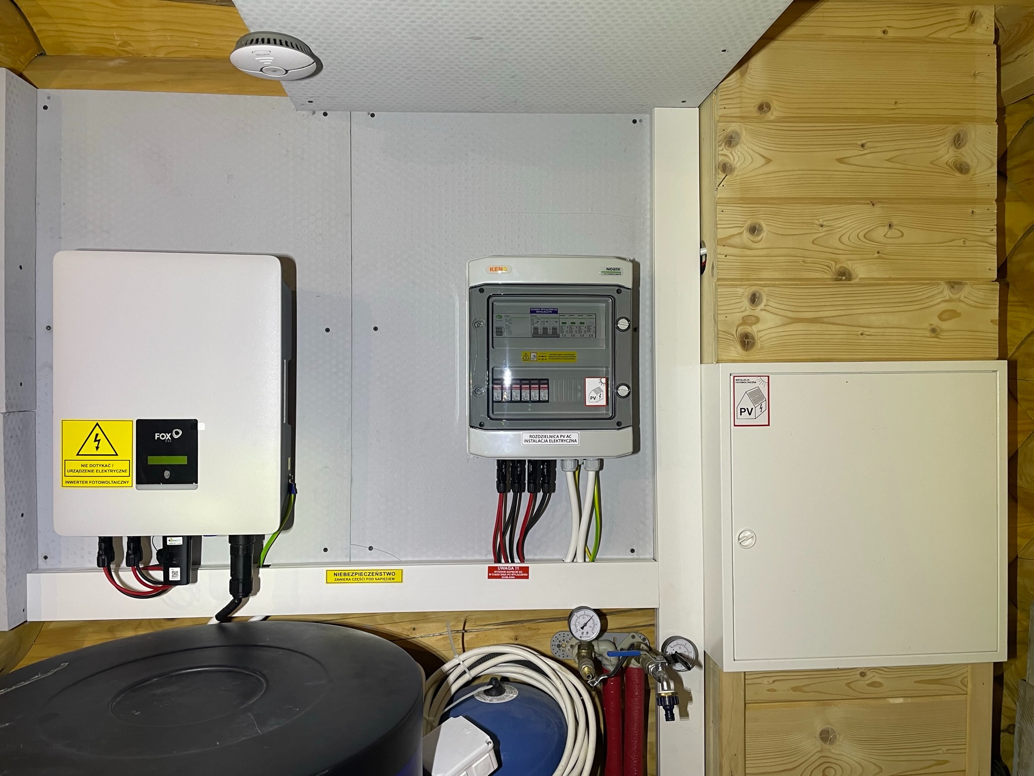 Inwerter fotowoltaiczny Fox ESS, rozdzielnica PV AC Noark i biała skrzynka instalacyjna na tle boazerii i płyty kartonowo-gipsowej, widoczne przewody i osprzęt instalacyjny.