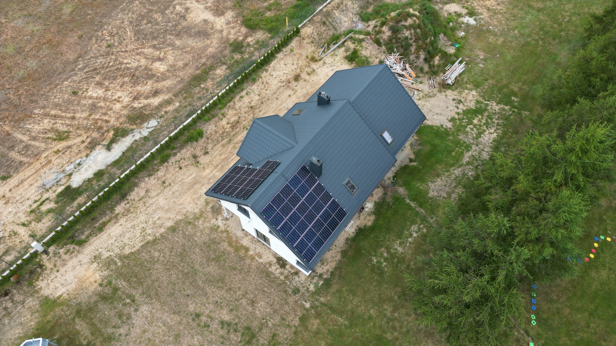 Widok z drona na dom jednorodzinny z panelami słonecznymi na dachu, otoczony terenem w trakcie zagospodarowywania i zielenią.